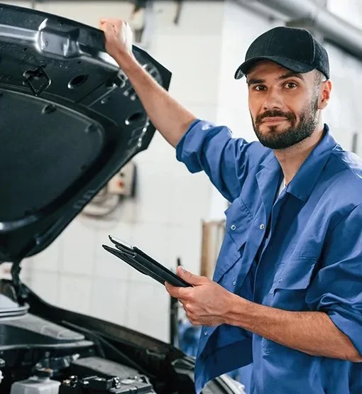 A mechanic inspecting a car engine with a clipboard in hand, wearing a blue uniform and black cap in an auto repair shop.