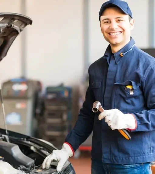 A smiling mechanic working on a car engine in a garage, holding a wrench and wearing gloves and a blue uniform.