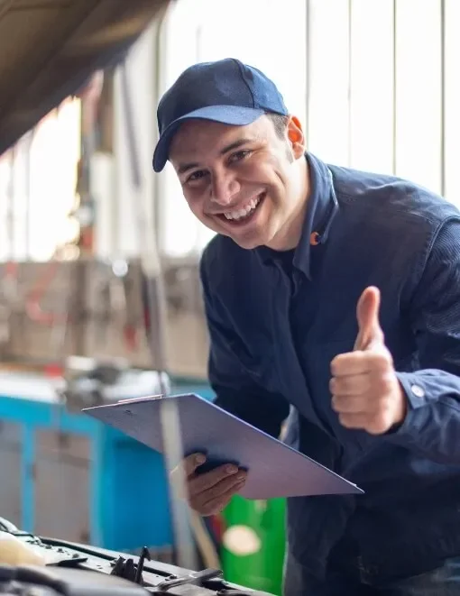 A smiling man wearing a blue uniform and cap, giving a thumbs-up while inspecting a car engine in an automotive repair shop.