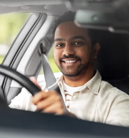 A smiling man driving a car, holding the steering wheel with one hand, with a seatbelt on.