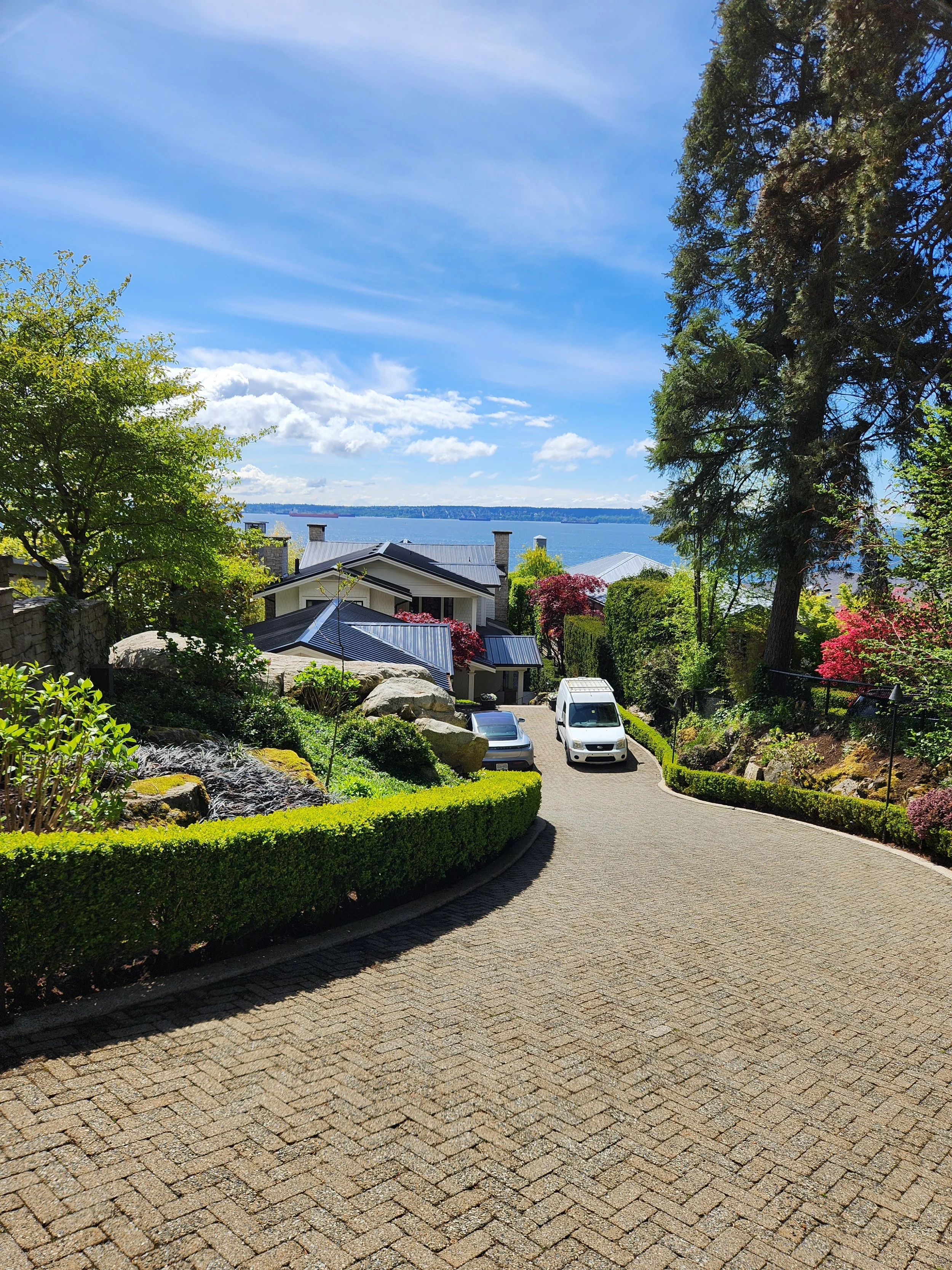 A paved residential driveway leading down to a house near a large body of water, with trees and colorful bushes on either side, under a bright blue sky with clouds.
