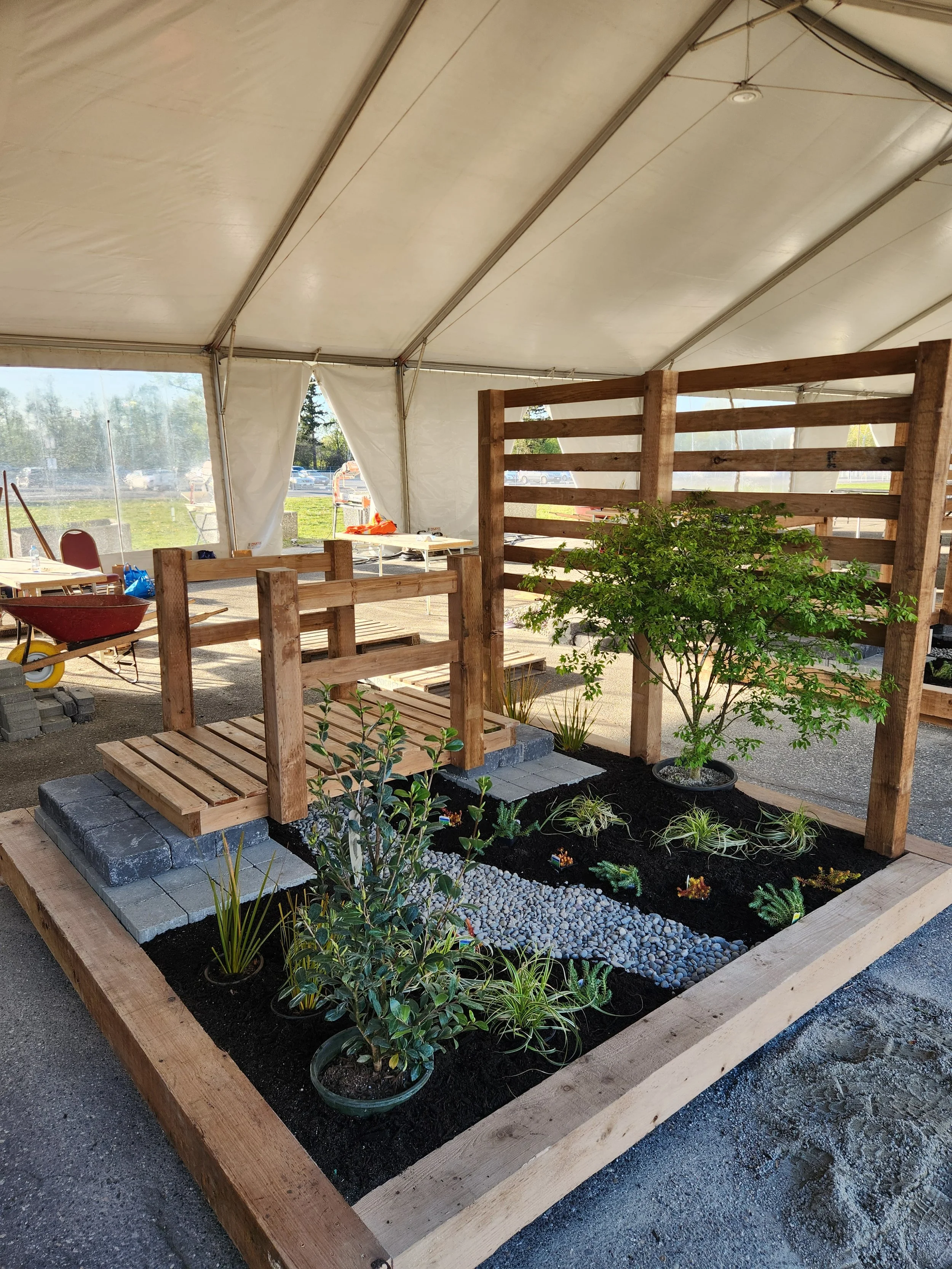 A small garden with potted plants and young trees, enclosed by a wooden rail, under a white tent.