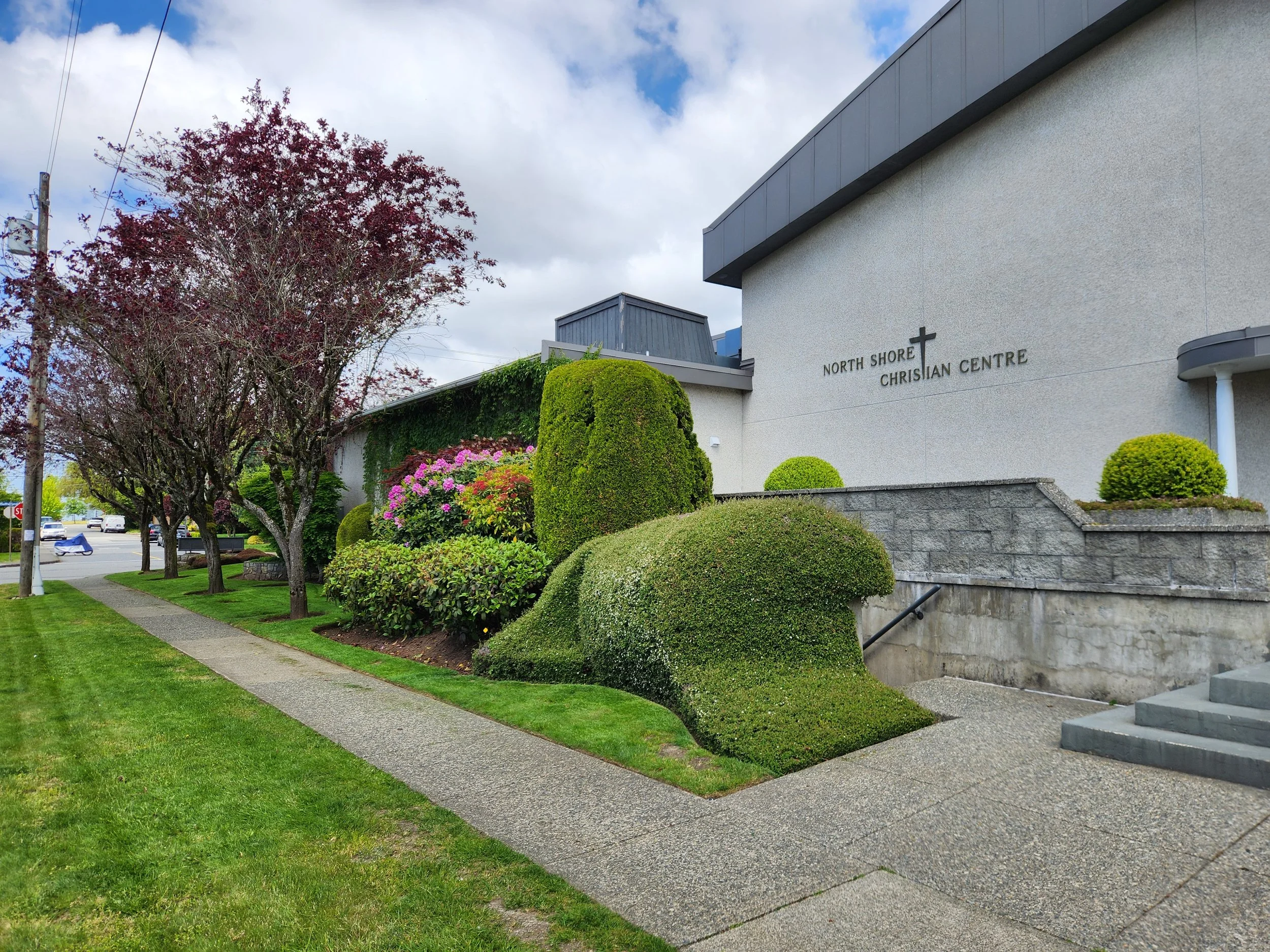 Exterior view of North Shore Christian Centre with manicured bushes, trees, sidewalk, and a street in the background.