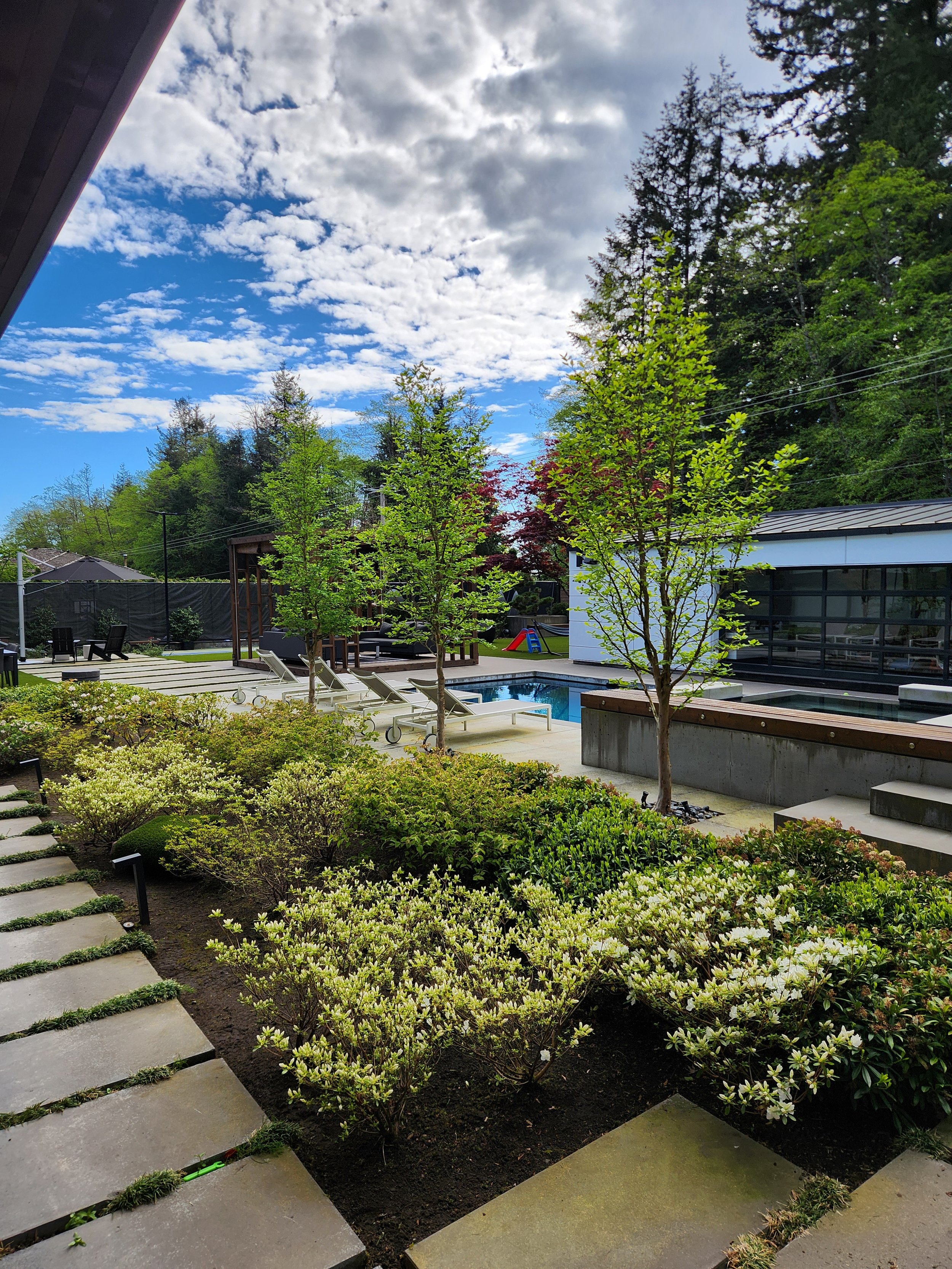 A backyard view showing a landscaped garden with shrubs and small trees, a patio with lounge chairs, a swimming pool, and a modern building with large windows, under a partly cloudy sky.