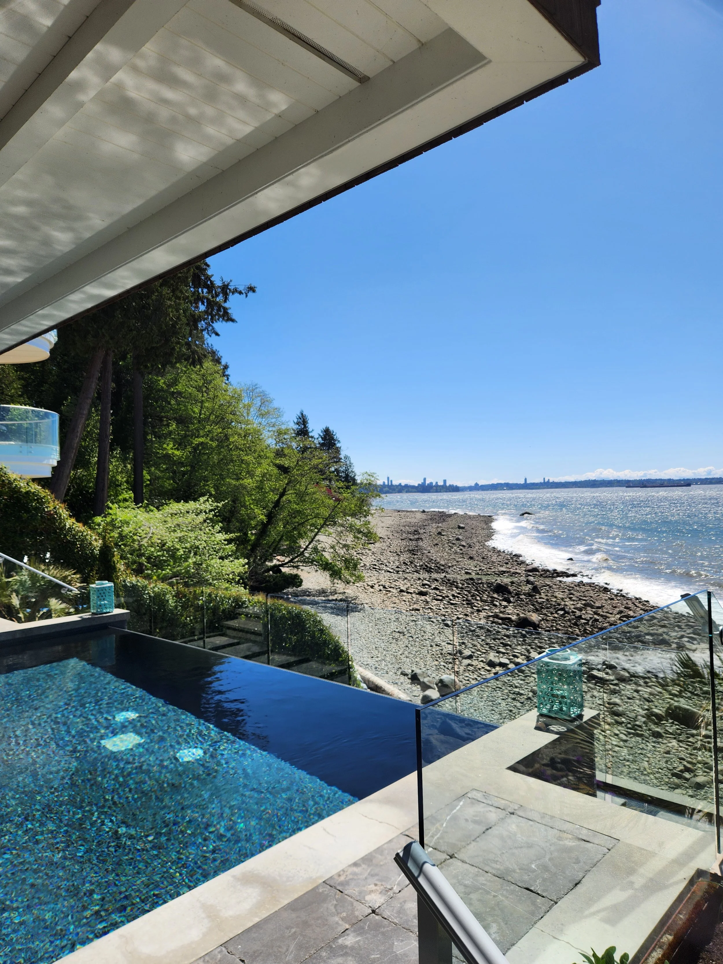 View of a private outdoor infinity pool overlooking a rocky beach and water, with trees and city skyline in the distance under a clear blue sky.