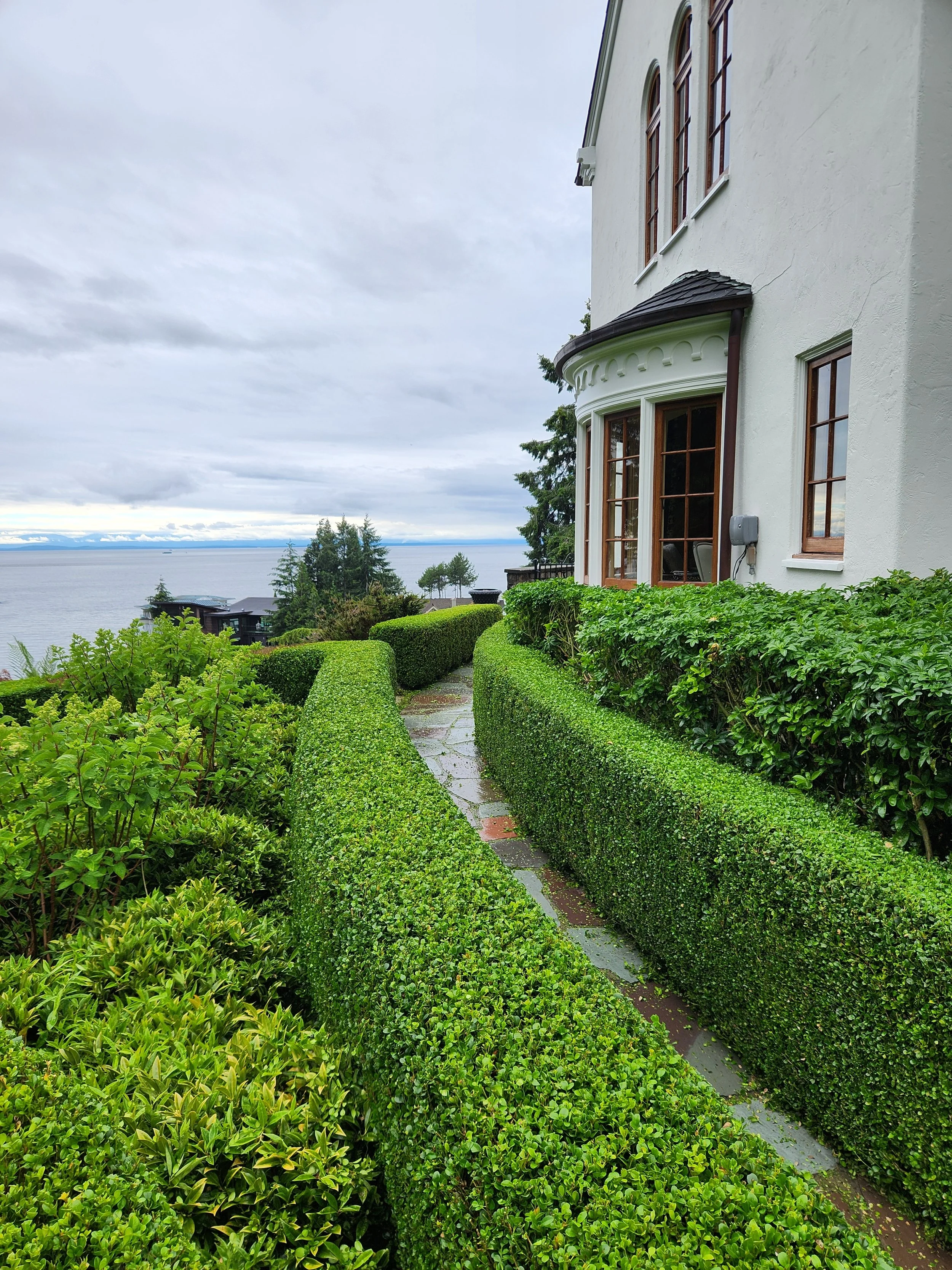 Pathway running alongside a large white house with brown trim, surrounded by neatly trimmed green bushes and trees, overlooking a body of water under a cloudy sky.