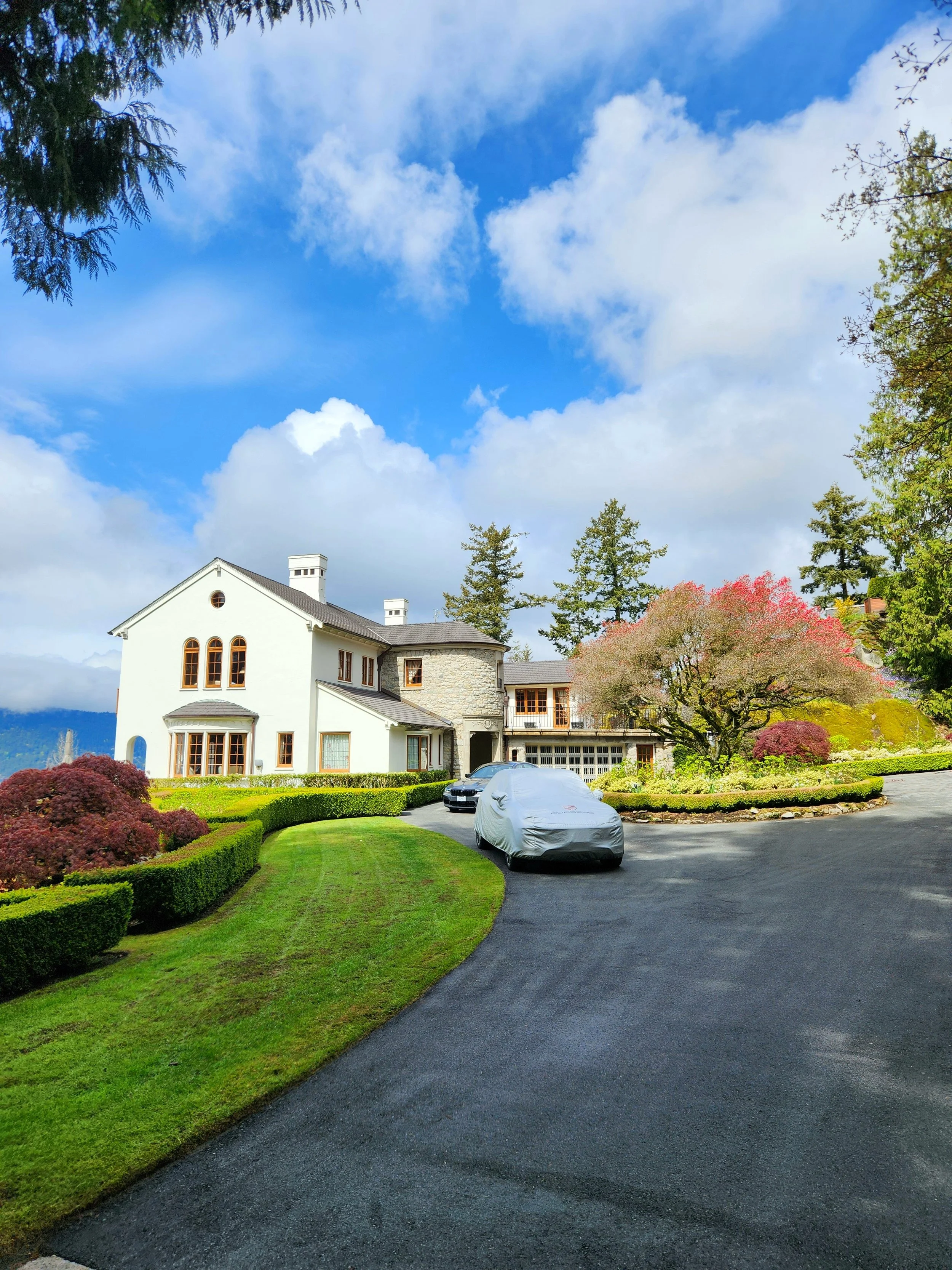 Large house surrounded by lush green landscaping, colorful trees, and a winding driveway with covered cars, under a partly cloudy blue sky.