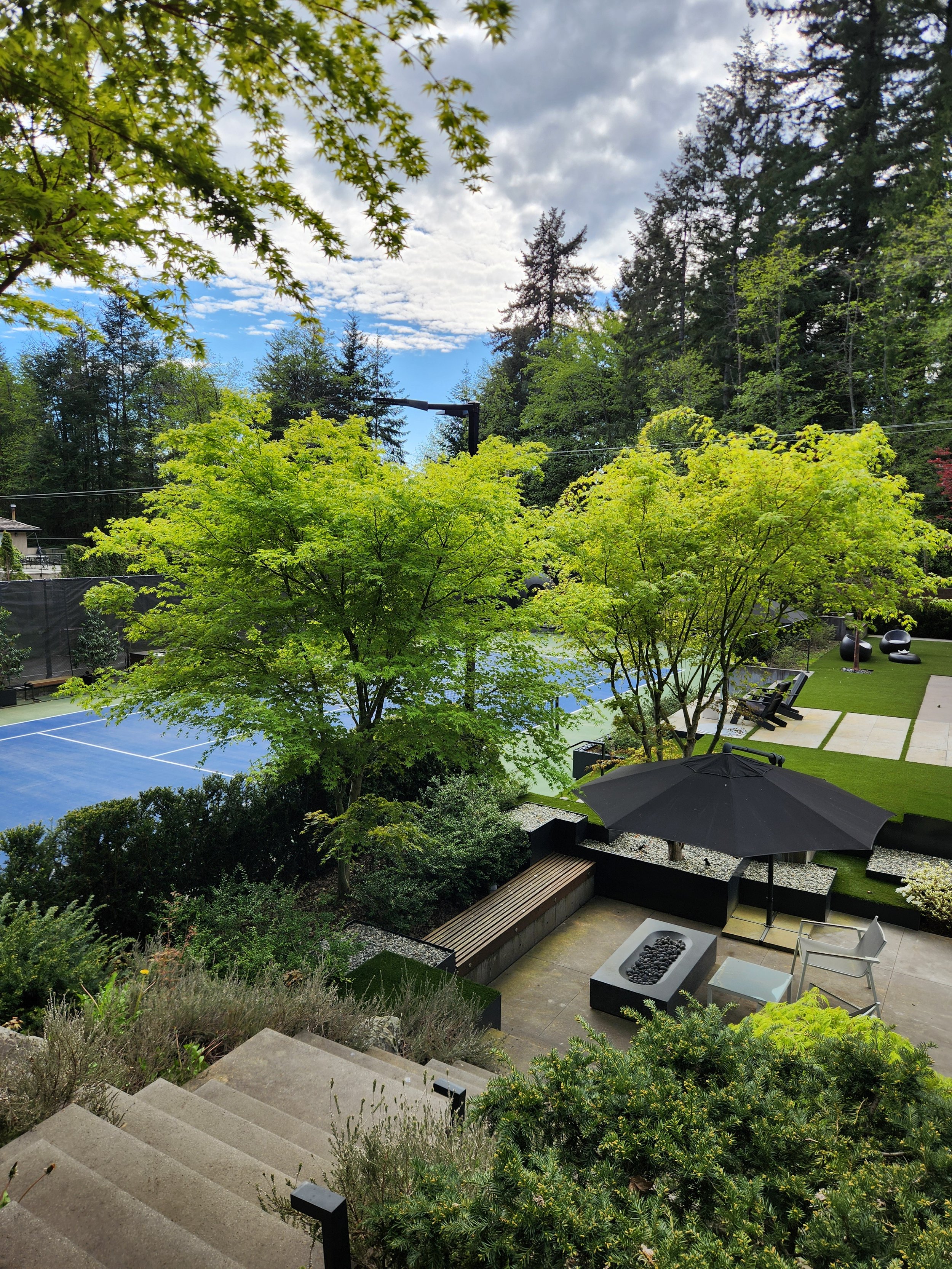 A backyard with a tennis court, lush green trees, a patio area with a black umbrella, chairs, a fire pit, and a well-maintained lawn surrounded by tall trees and a forested area.