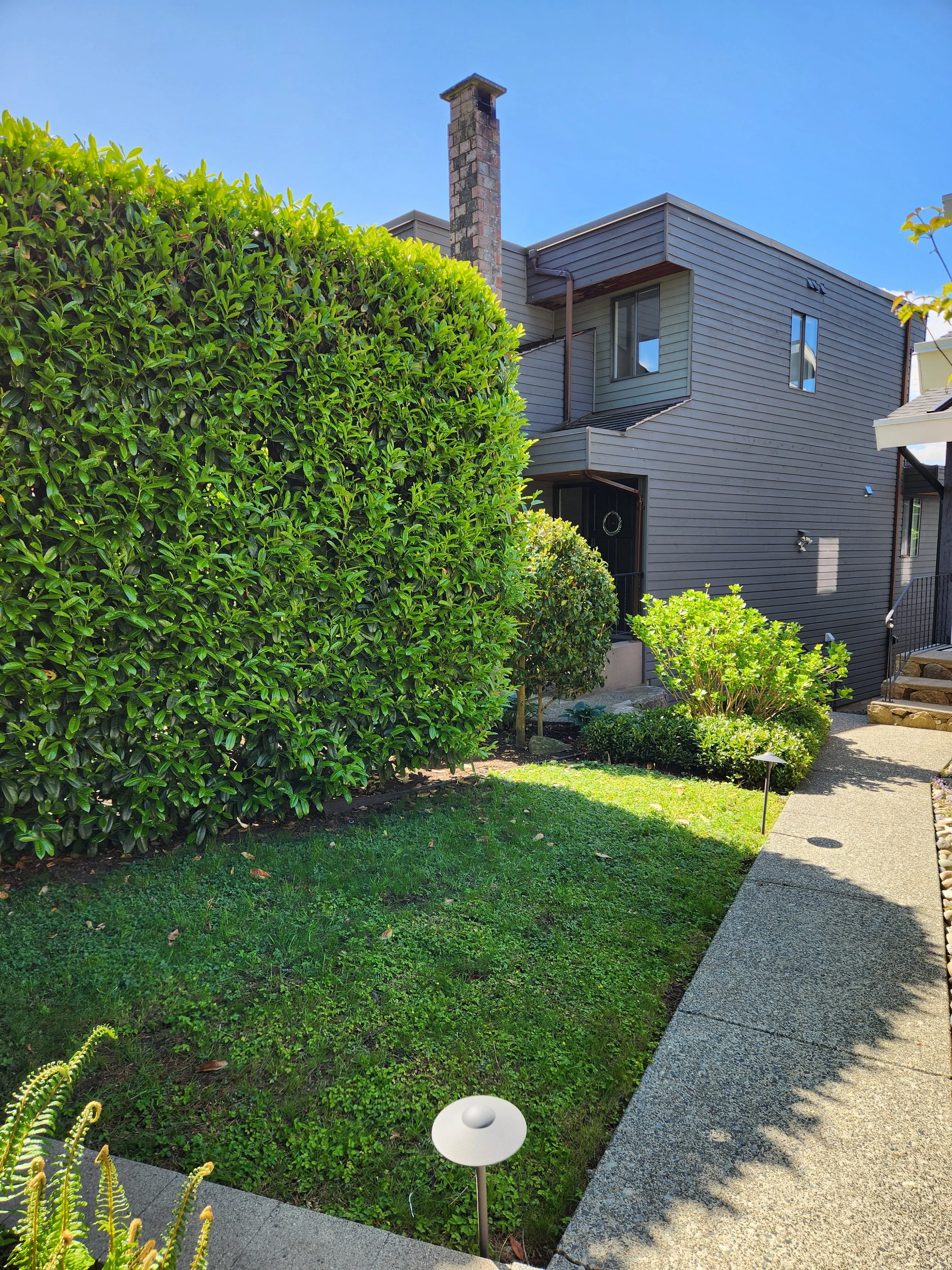 A well-maintained garden with lush green shrubs and a grass lawn, adjacent to a grey multi-story house with dark siding and a brick chimney, under a clear blue sky.