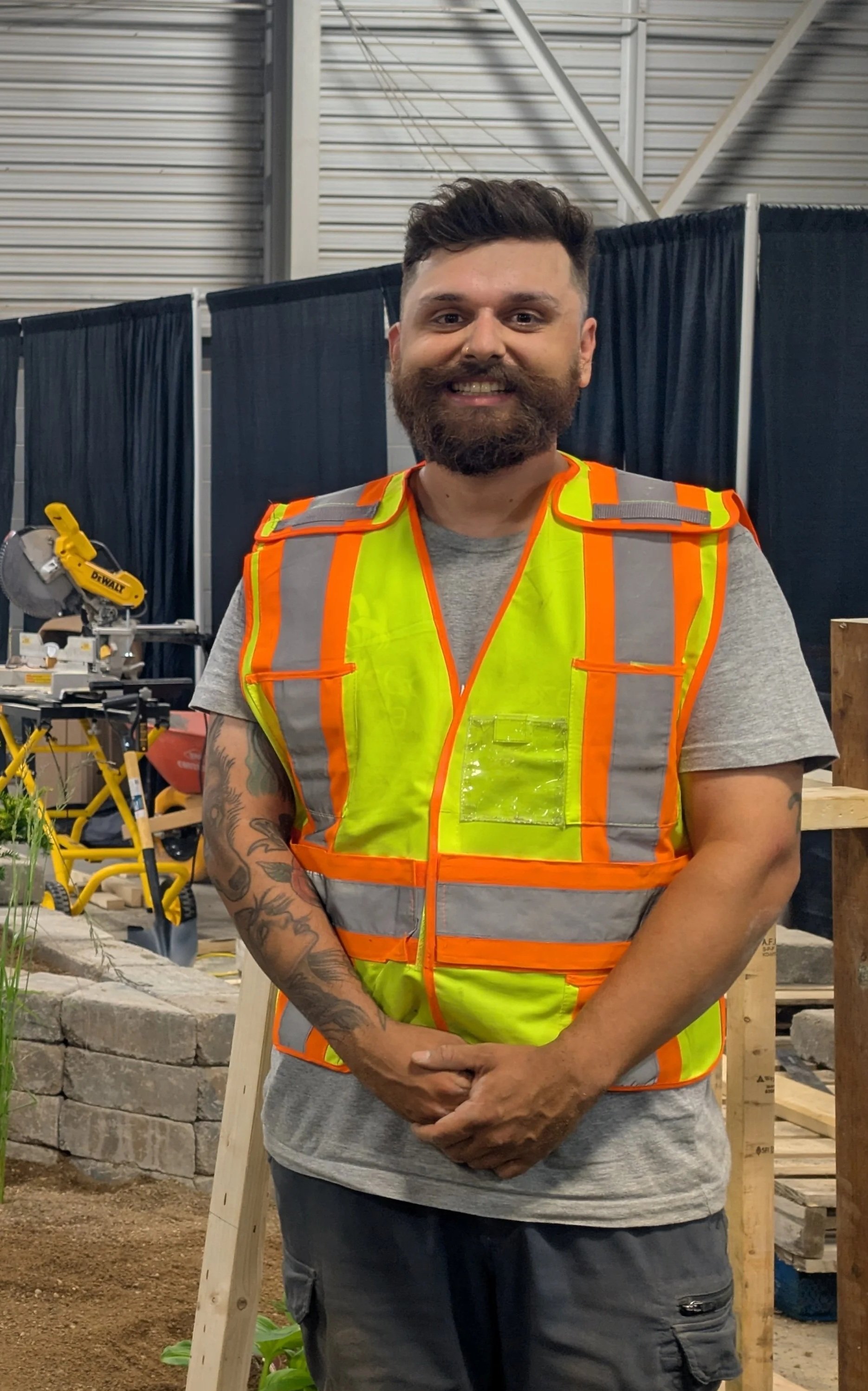 Man with a beard and tattoos wearing a gray T-shirt and a bright yellow safety vest with orange reflective stripes, standing in a construction or woodworking area with tools and wooden structures in the background.
