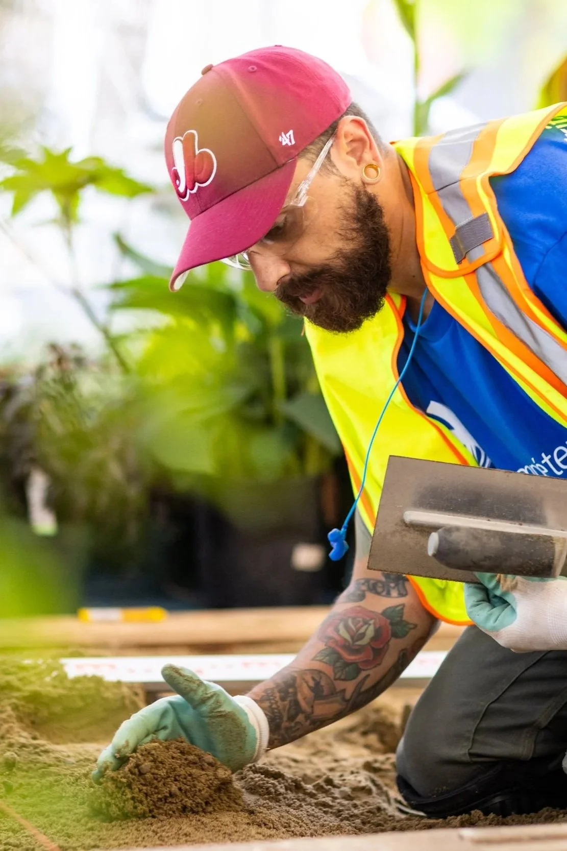 Man wearing safety glasses, a red cap, yellow safety vest, and gloves, using a trowel to work with soil or sand, with plants in the background.