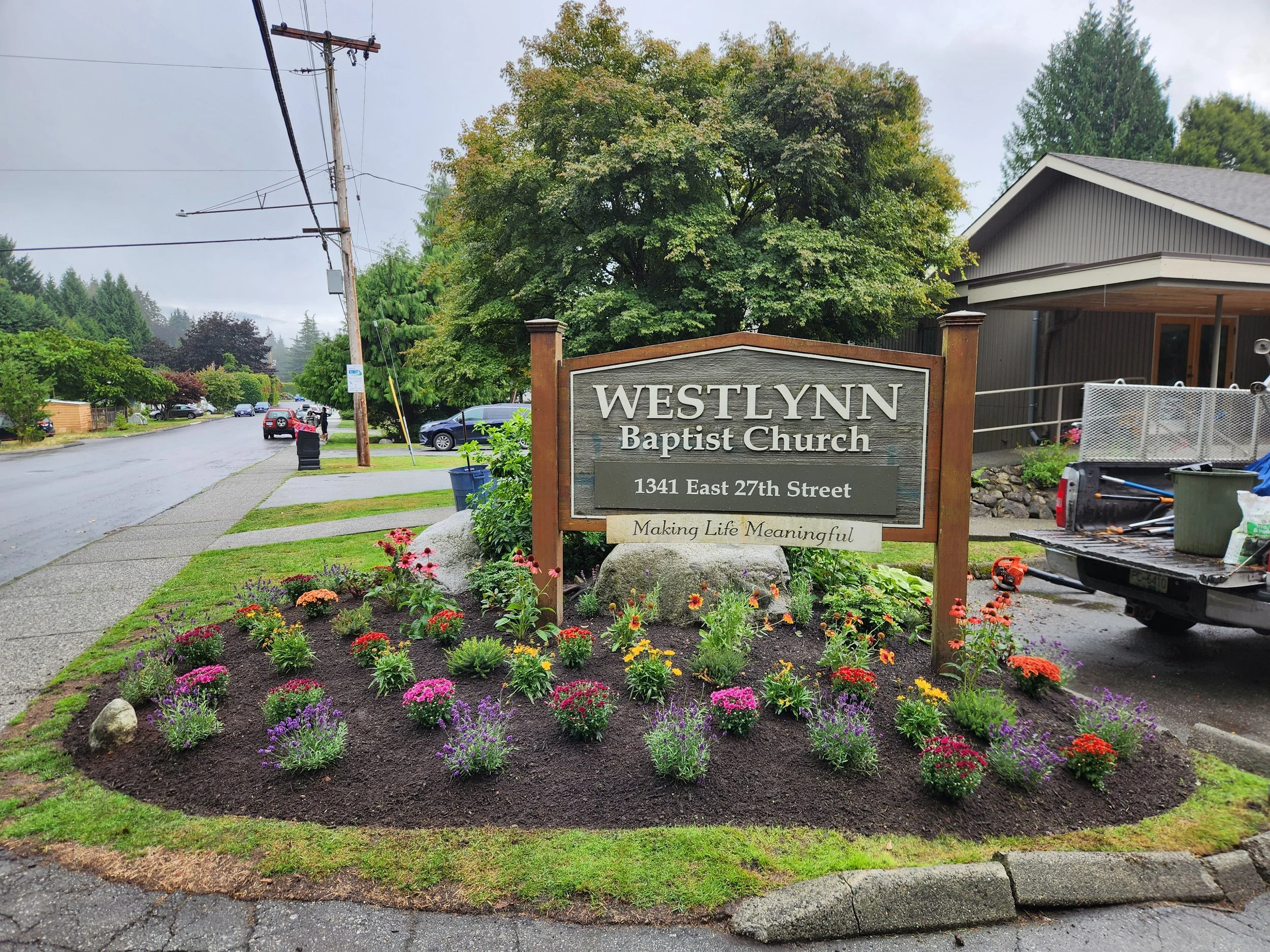 Street view of Westlynn Baptist Church sign with flowers in front, a house to the right, and wet sidewalk on rainy day.
