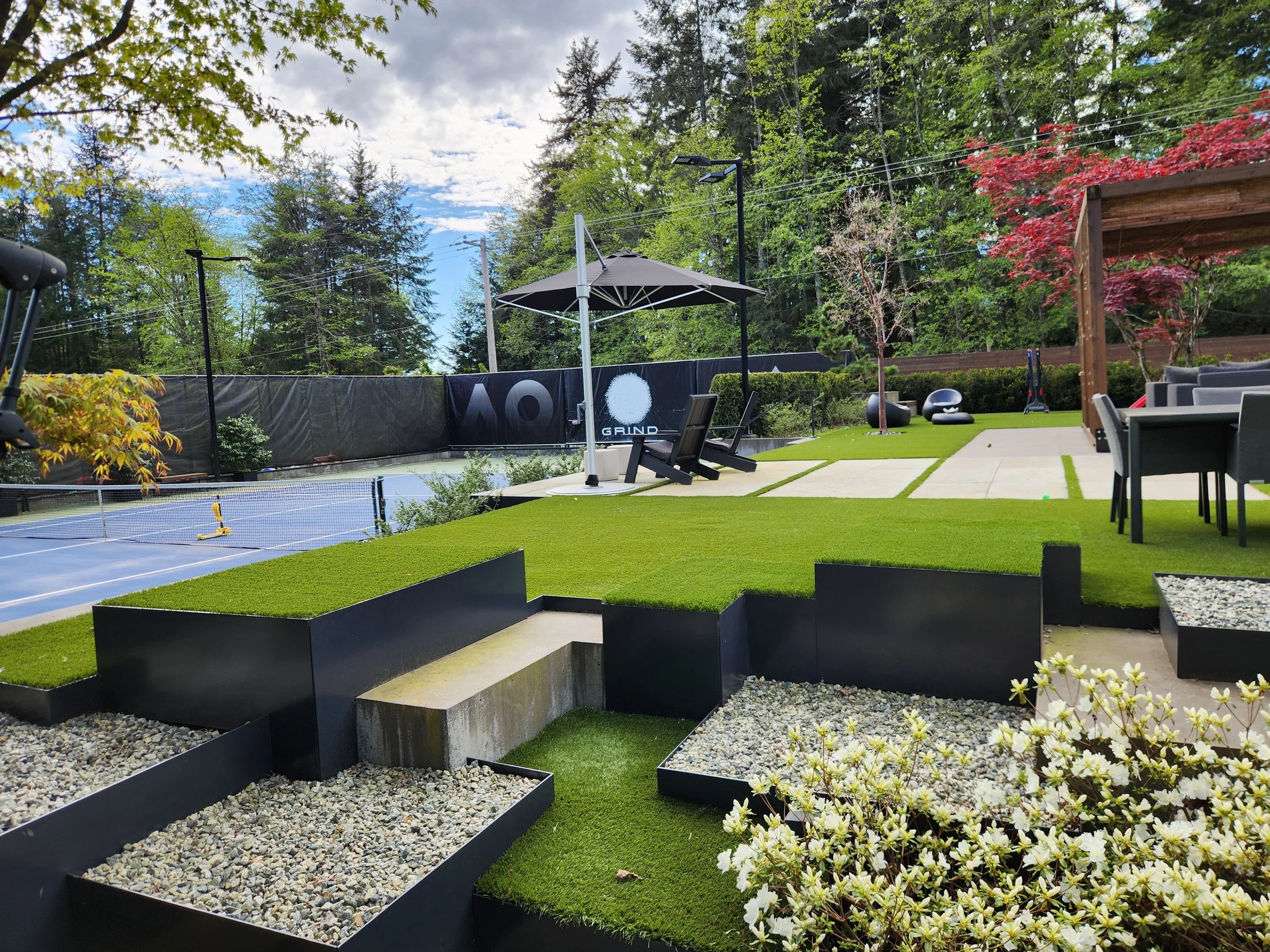 A backyard with a tennis court, outdoor seating, lounge chairs, and landscaped plants, surrounded by tall trees and a partly cloudy sky.