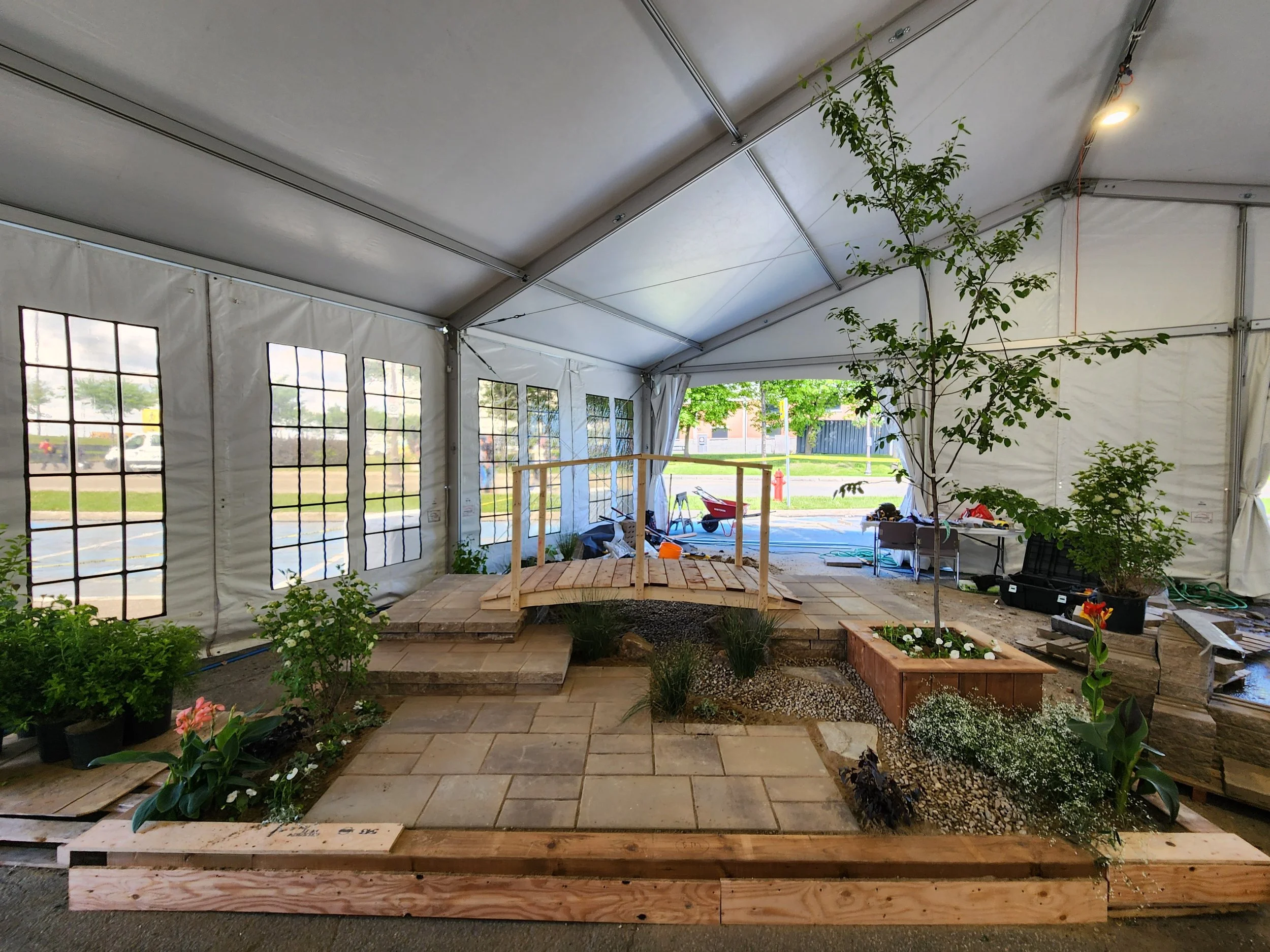 Indoor garden space with potted plants, a small wooden bridge, stone pathway, trees, and gardening supplies inside a large white tent.