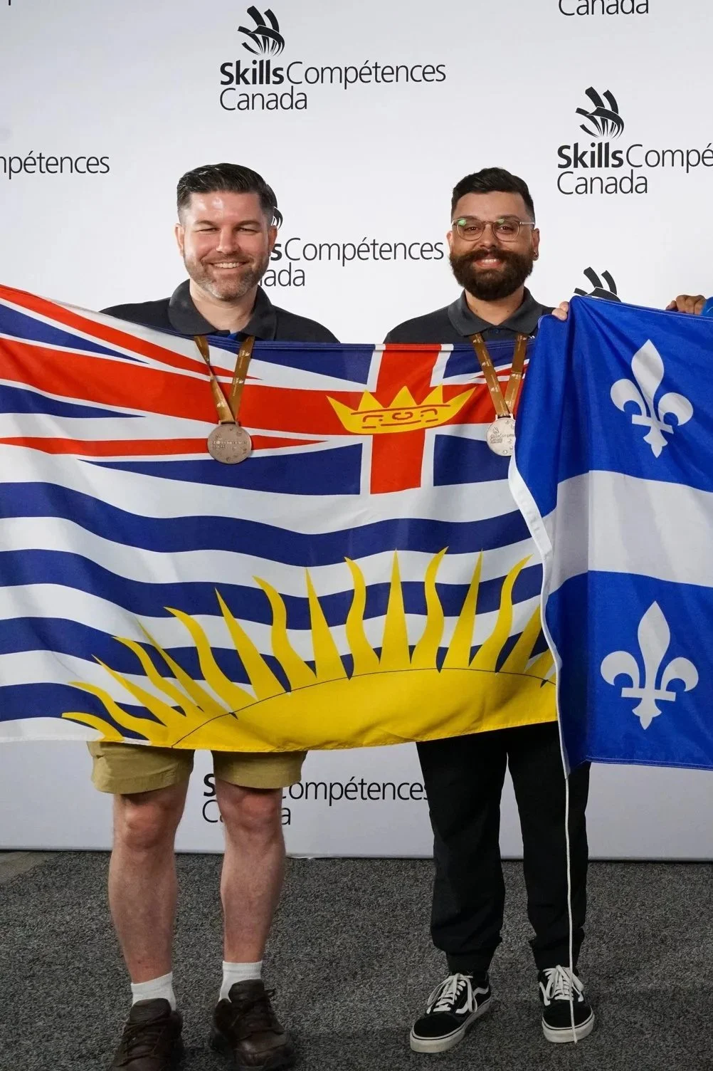 Two men standing side by side, holding flags and wearing medals, in front of a Skills Canada backdrop. One man holds a flag with a yellow sun and blue, red, and white stripes, while the other holds a blue and white flag with a fleur-de-lis. They are smiling, and the event appears to be a competition or awards ceremony.