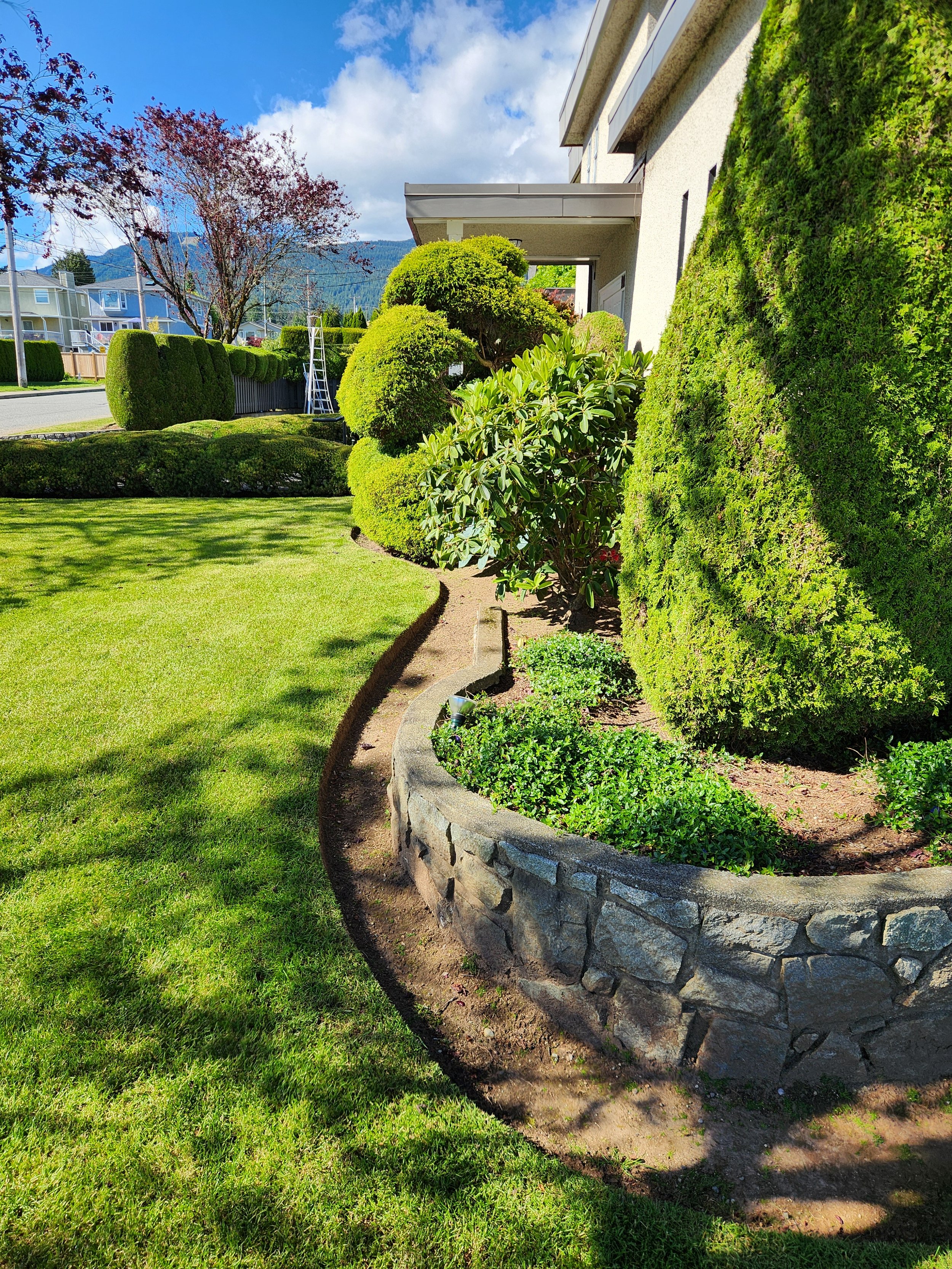 Well-maintained front yard with green grass, bushes, and trees, with a stone border separating the garden bed from the lawn, and a house in the background under a partly cloudy sky.