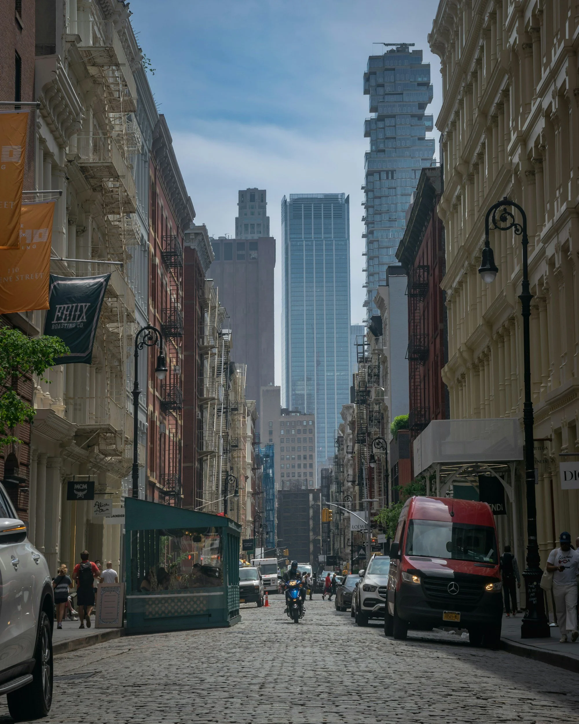 A city street scene in New York City with buildings and skyscrapers, including a unique stacked residential tower, cobblestone road, parked cars, a motorcycle, pedestrians, street lamps, and storefronts.