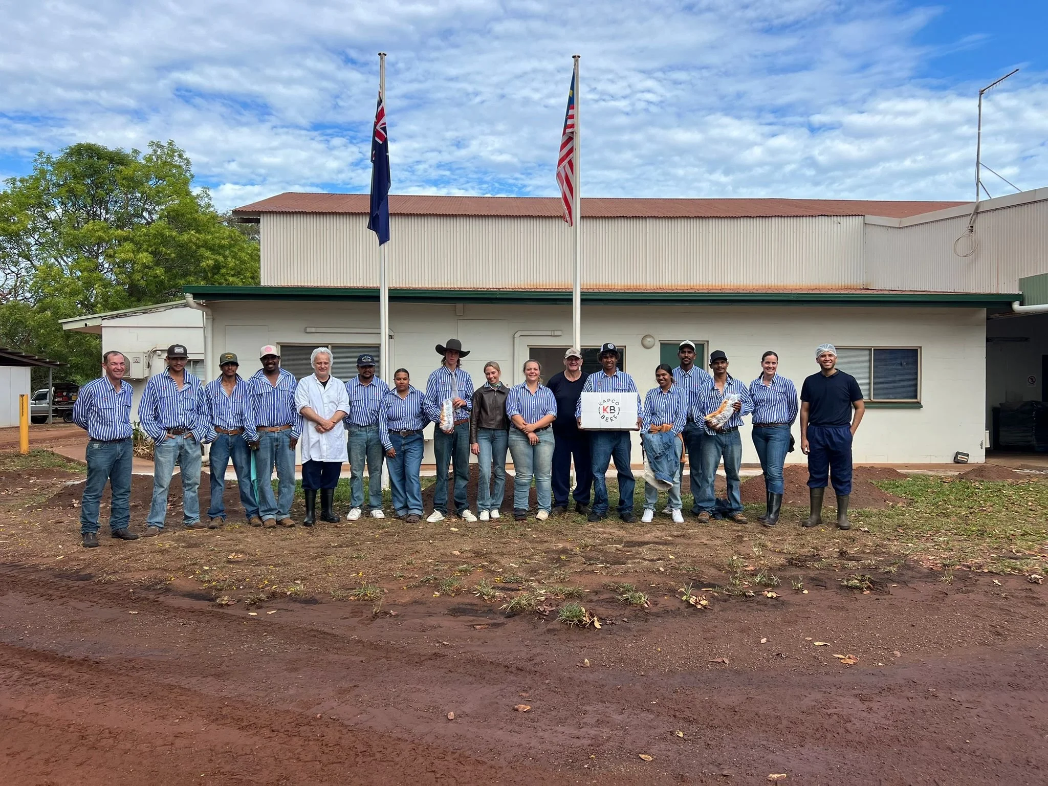 A group of people standing outdoors in front of a building with two flags on flagspoles, one with the Australian flag and the other with the United States flag, under a partly cloudy sky.