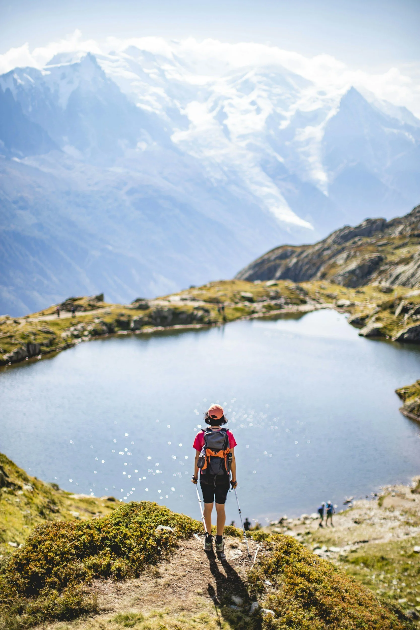A person with a backpack and hat hiking near a mountain lake with snow-capped peaks in the background.