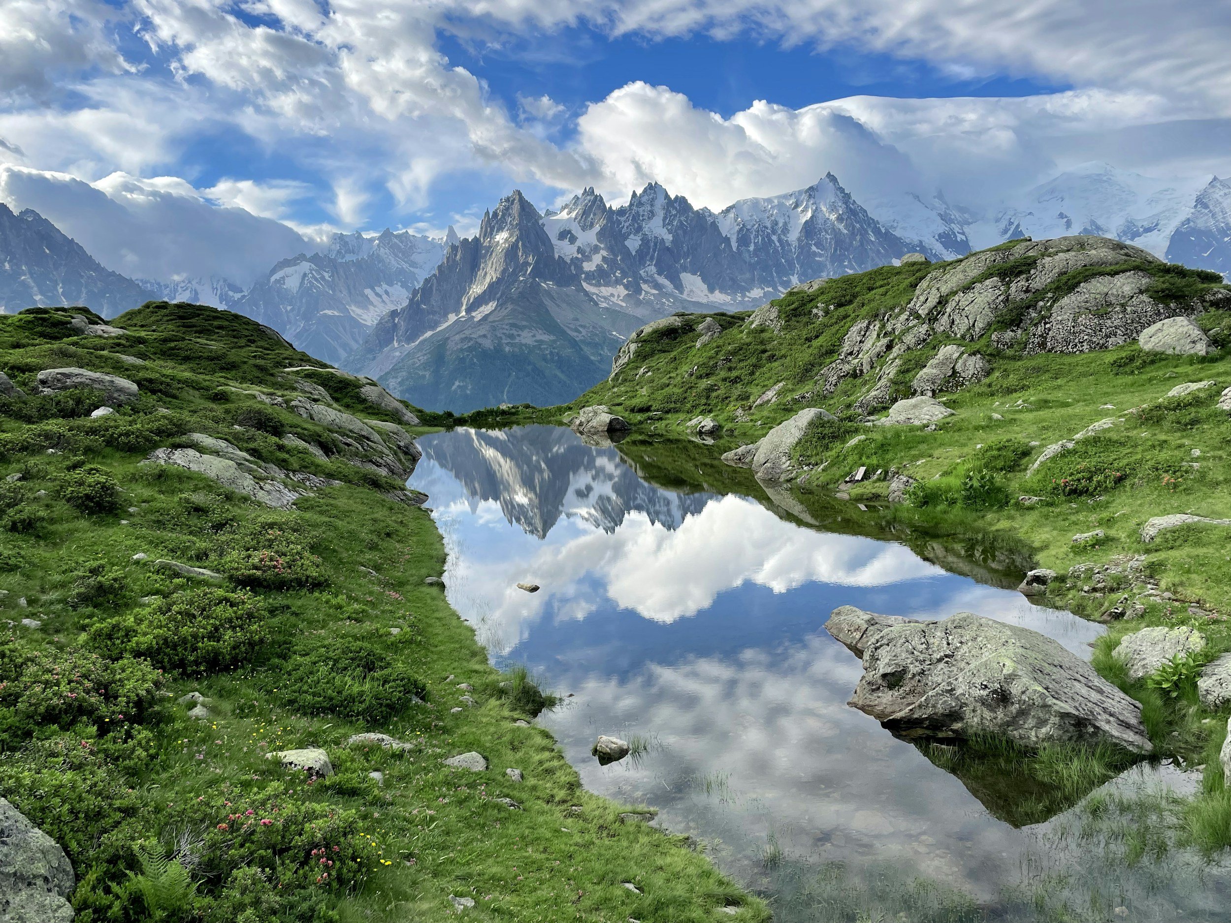Mountain landscape with snow-capped peaks, lush green hills, and a reflecting lake with clouds and sky mirrored in the water.