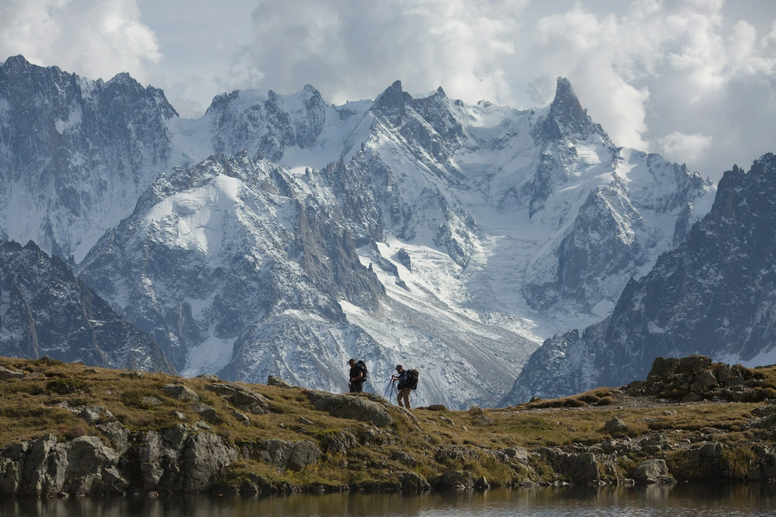 Two hikers with backpacks stand on a grassy patch near a small body of water, with snow-covered mountains in the background.