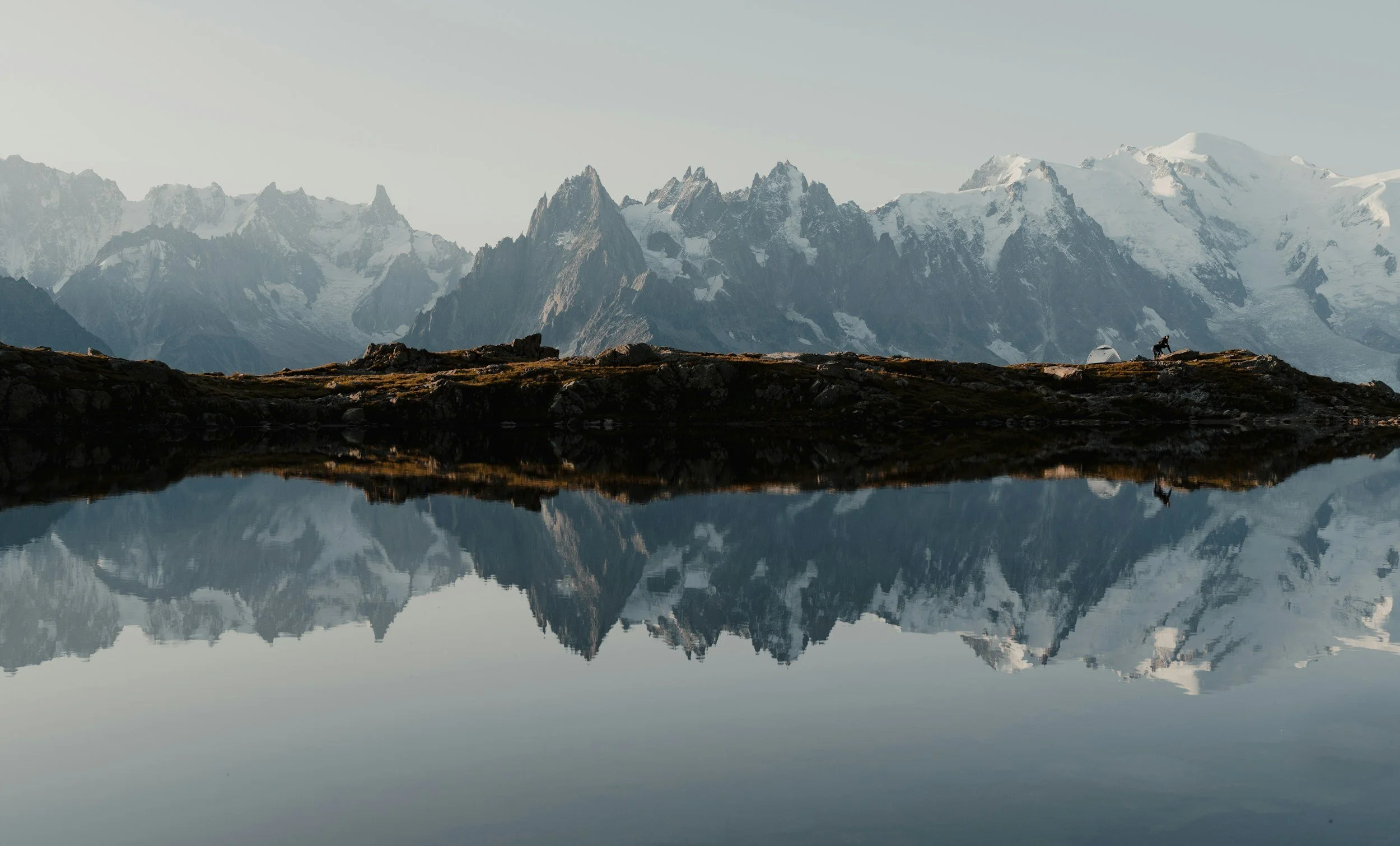 Reflection of snow-capped mountains and rocky terrain on a calm lake, with a person and a tent on the shoreline.