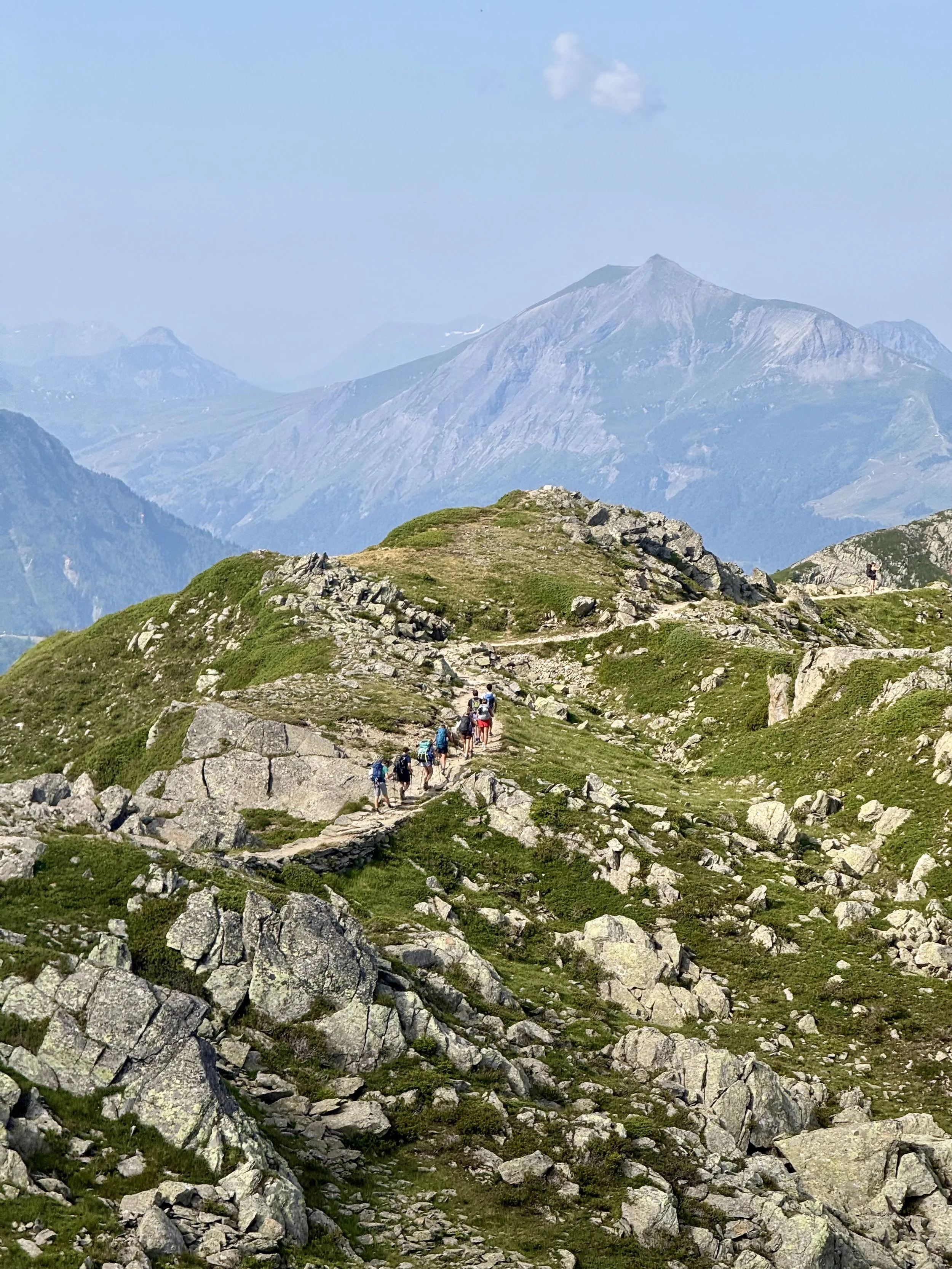 Group of trail hikers walking through rocky mountain landscape with green vegetation, overlooking distant mountain peaks under a blue sky.