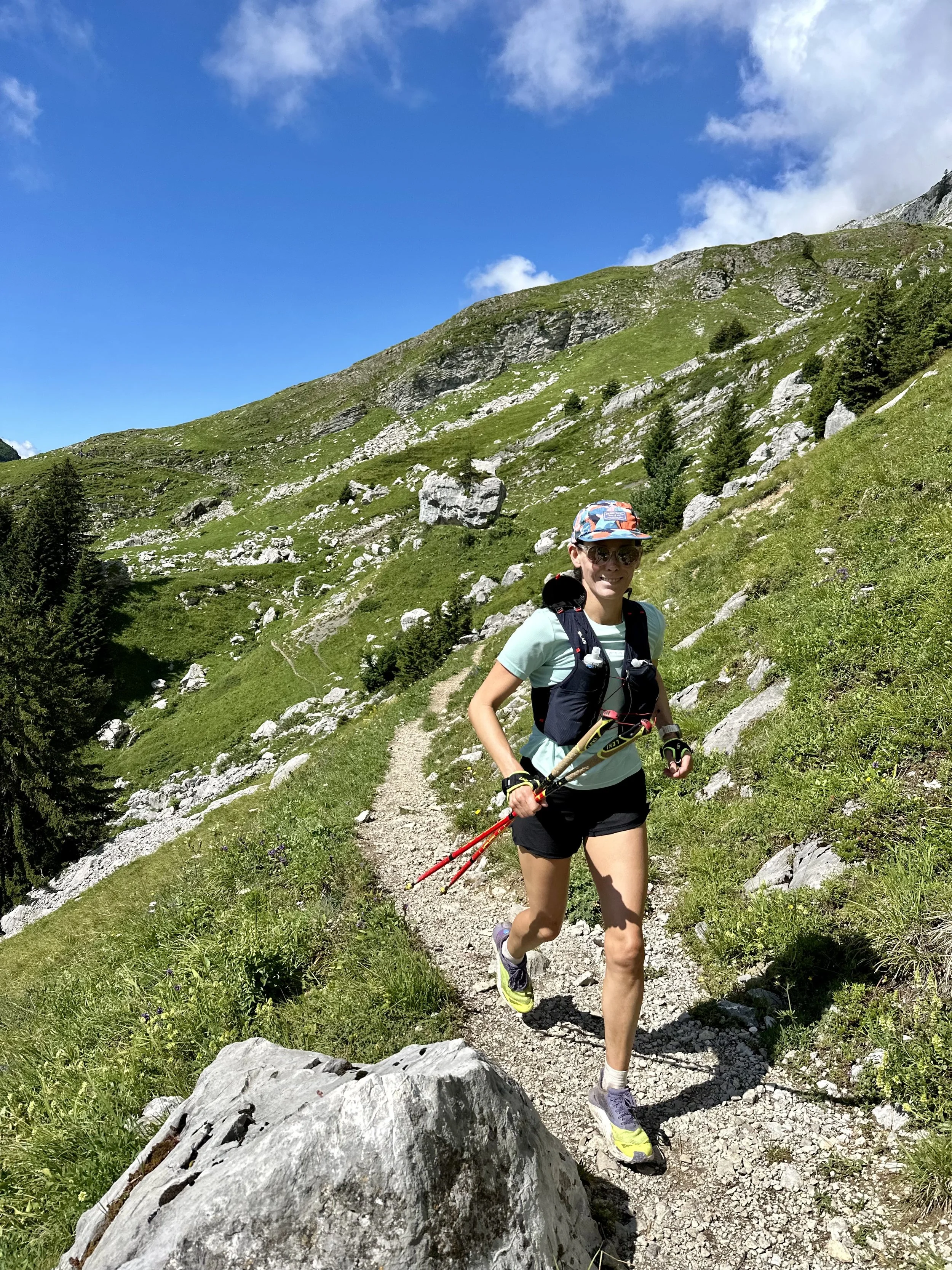 A woman trail running on a narrow mountain path with green hills, rocks, and trees under a partly cloudy sky.