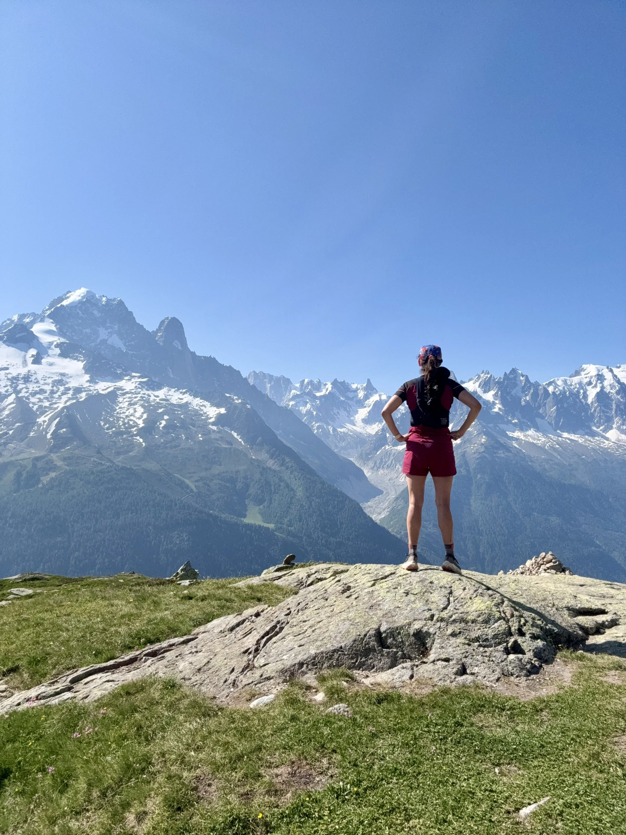 A woman stands with hands on hips on a large rock, overlooking snow-capped mountains under a clear blue sky.