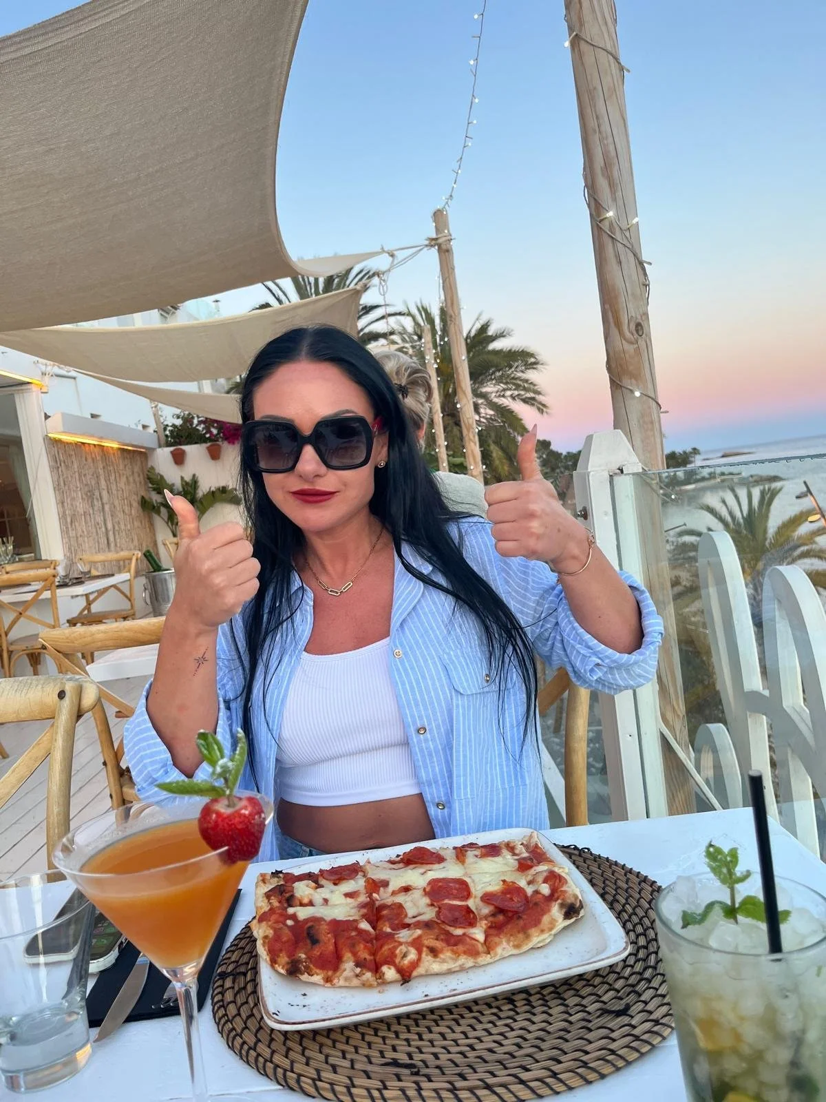 A woman with black hair wearing sunglasses and a striped blue shirt gives two thumbs up at a seaside restaurant table with a pizza and cocktails, during sunset with palm trees in the background.