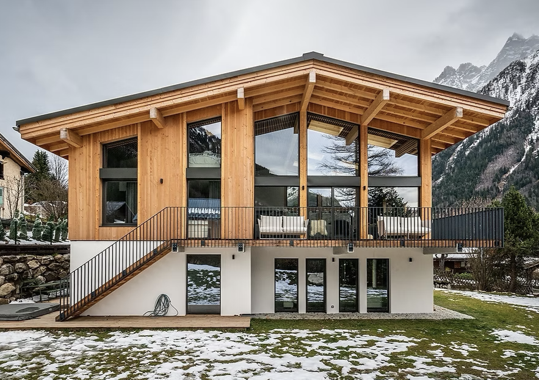 Modern wooden house with large windows and balcony, snow-covered ground, mountainous background, and partly cloudy sky.