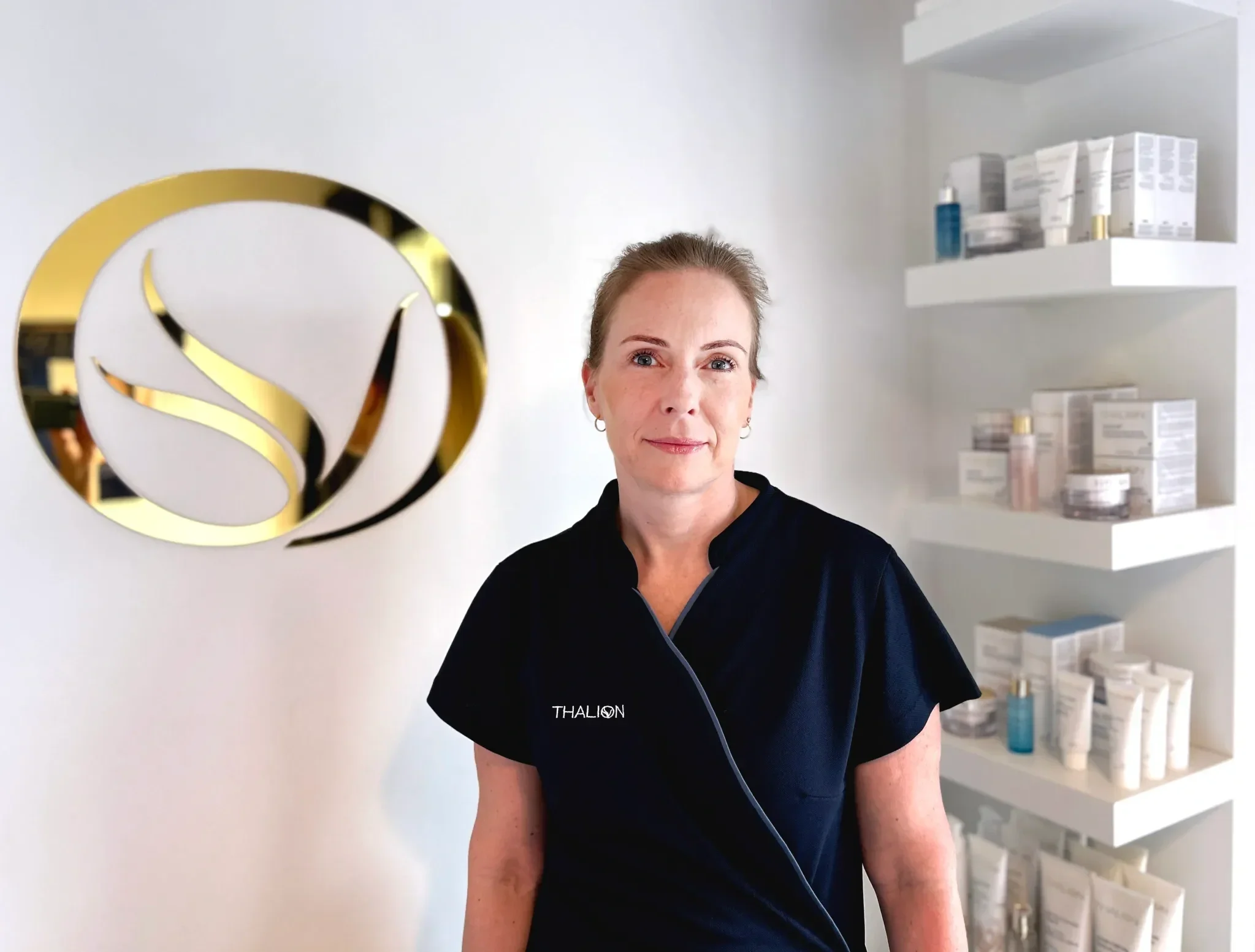 A woman standing in a spa or skincare clinic, wearing a navy uniform with the brand name 'Thalion' embroidered, smiling softly, with shelves of skincare products behind her and a decorative gold and white logo on the wall.