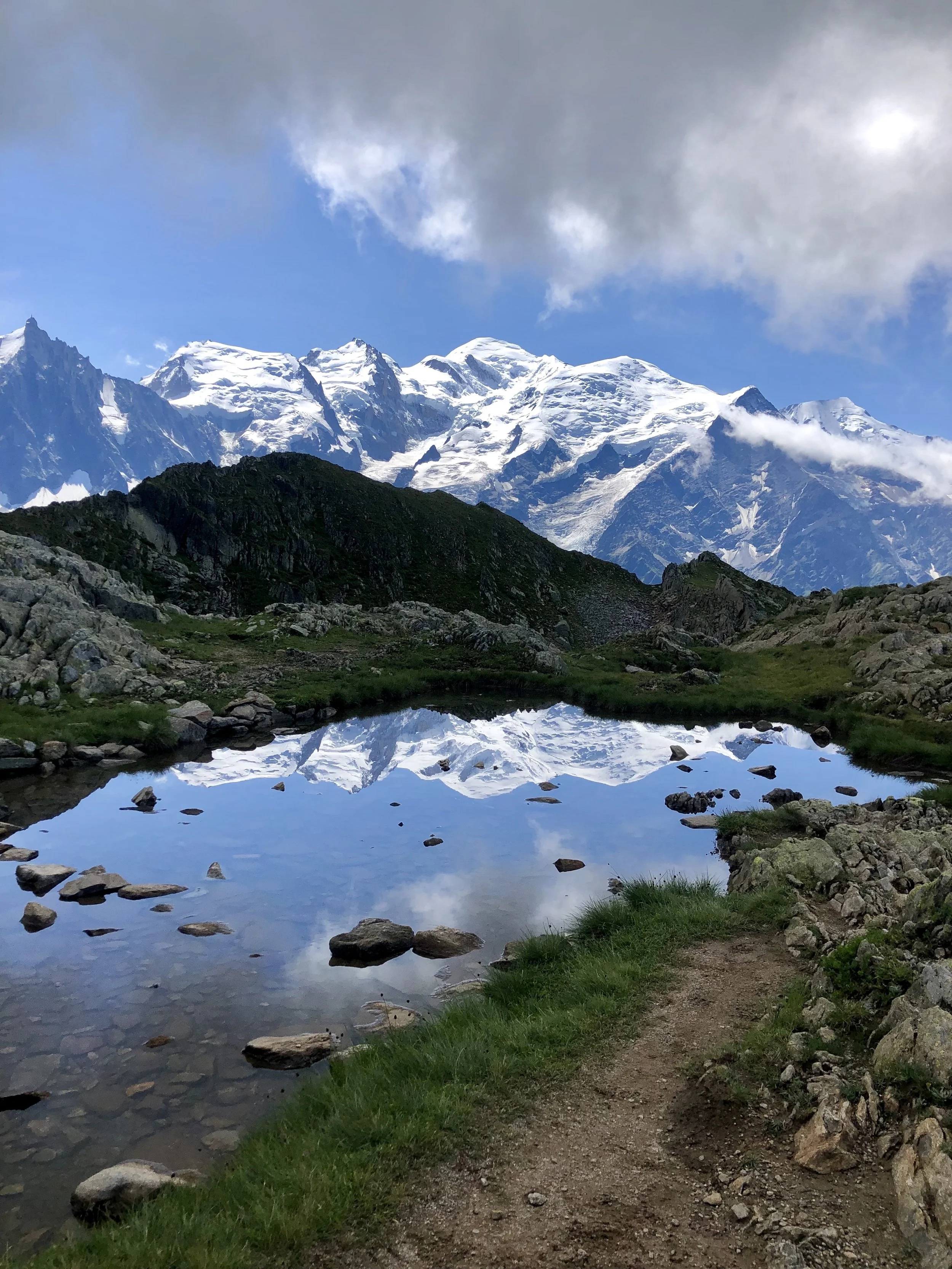 A hiking trail beside a small pond with still water, reflecting snow-covered mountain peaks under a partly cloudy sky.