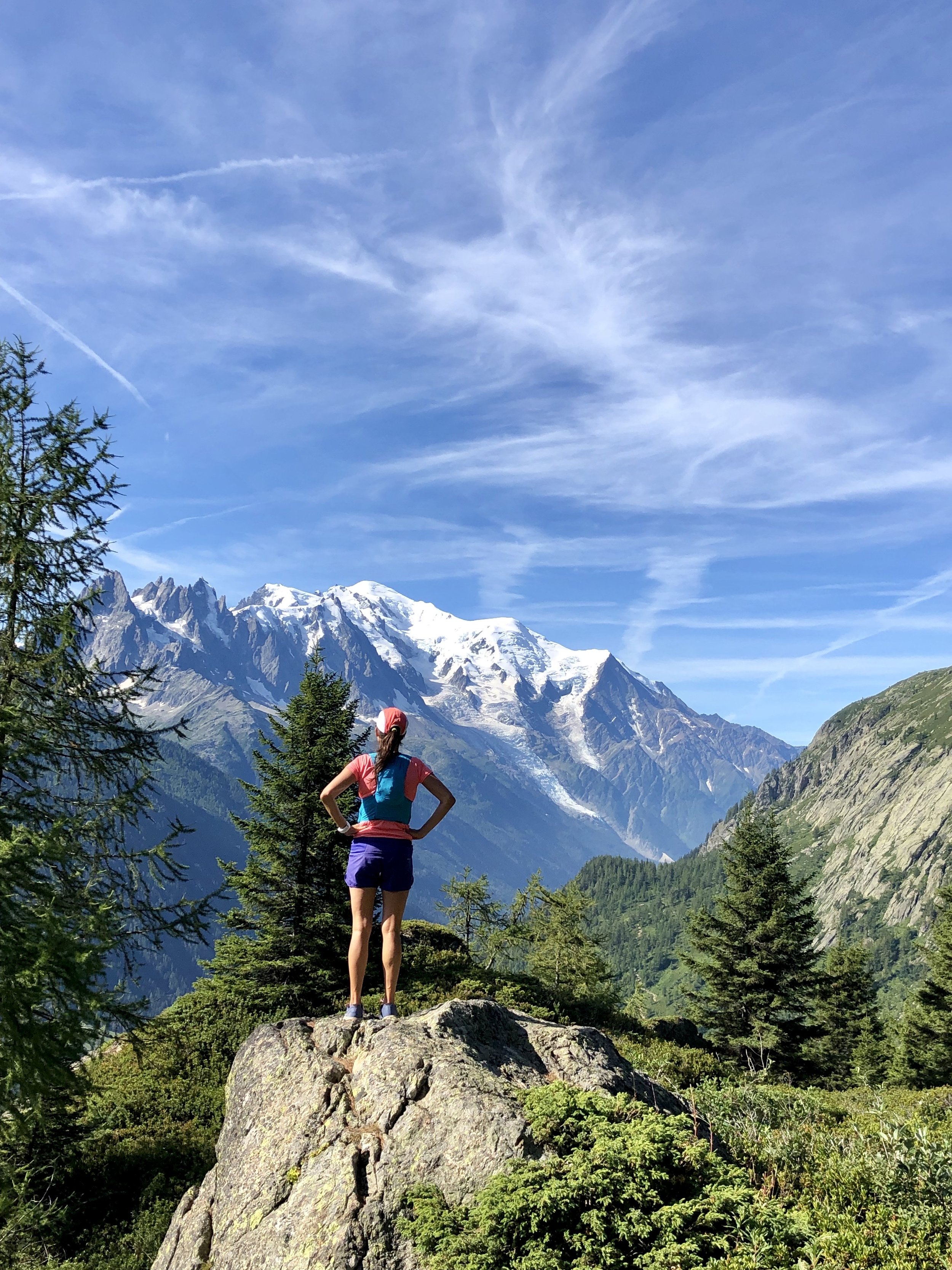 A woman standing on a large rock in a mountainous landscape with snow-capped peaks, green forest, and a blue sky with wispy clouds.
