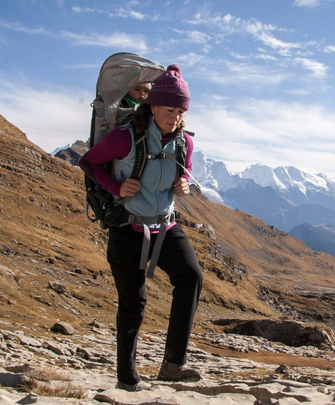 A woman hiking on a rocky trail with a child in a backpack carrier on her back, mountains in the background, under a partly cloudy sky.