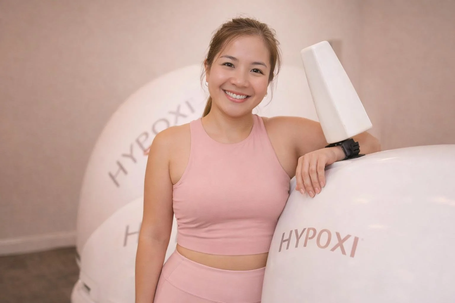 A smiling woman in pink workout clothes leaning on large white hypoxia training equipment in a fitness center.