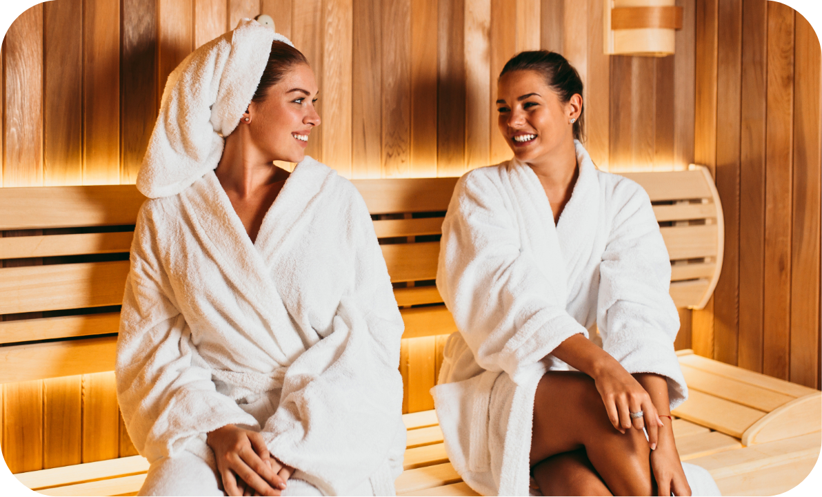 Two women in white bathrobes sitting on a bench inside a wooden sauna, smiling and enjoying their time.