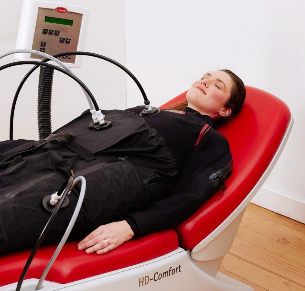 A woman lying on a medical bed connected to a hyperbaric oxygen chamber for therapy.