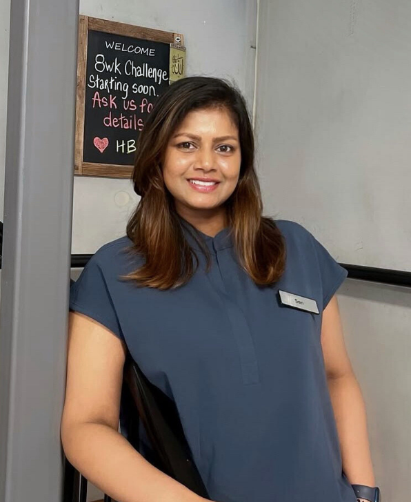 A woman with shoulder-length brown hair smiling in a gray uniform with a name tag, standing indoors near a wall and a black chalkboard with a welcome message and a challenge announcement.