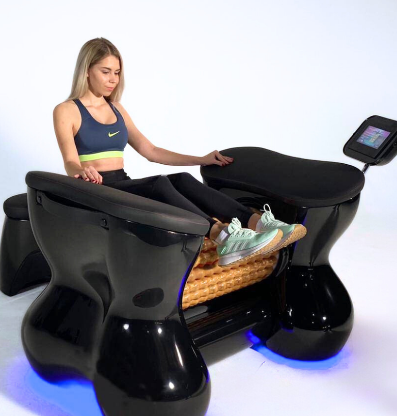A woman in workout clothes sitting in a futuristic in-home massage chair with a built-in footrest and digital screen.