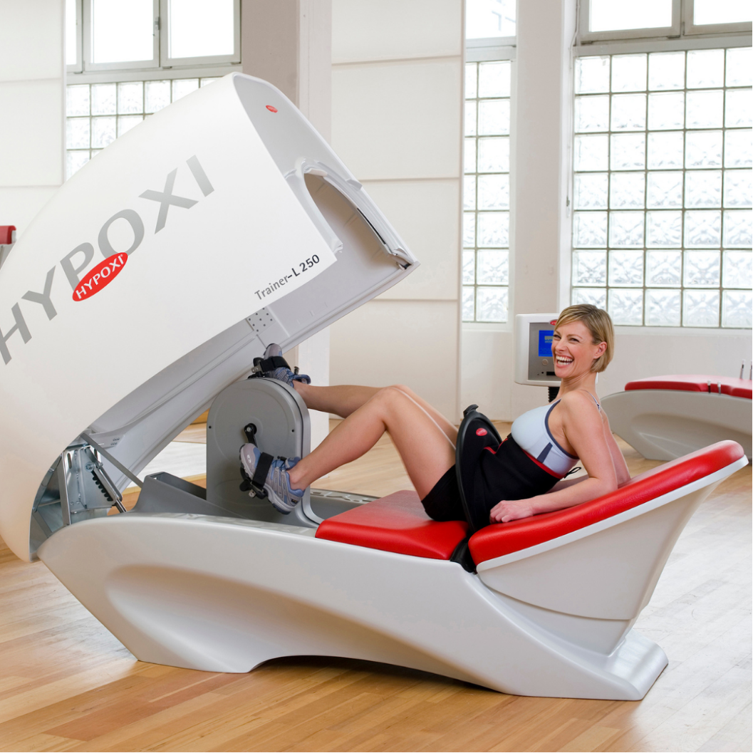 A woman smiling and sitting on a treadmill-like machine used for medical imaging, with her legs inside the scanner, in a well-lit room with large windows.