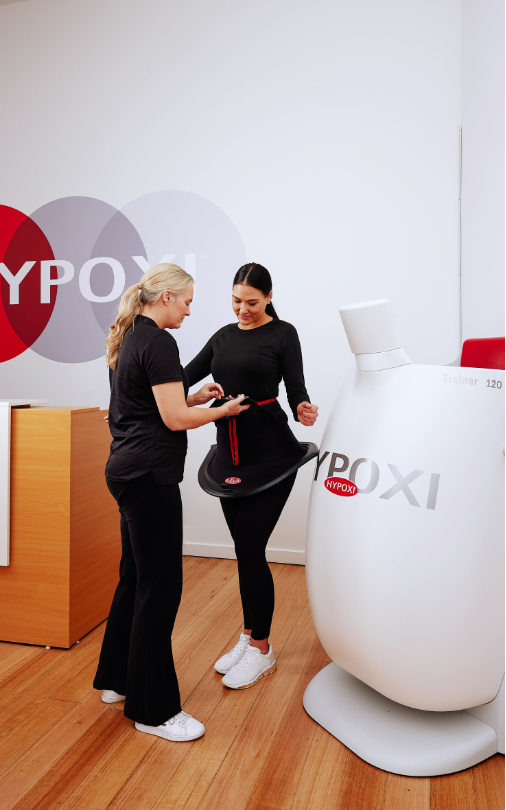 Two women interact at a Hypoxi booth with a large white machine in a modern room, one woman displaying a device to the other.