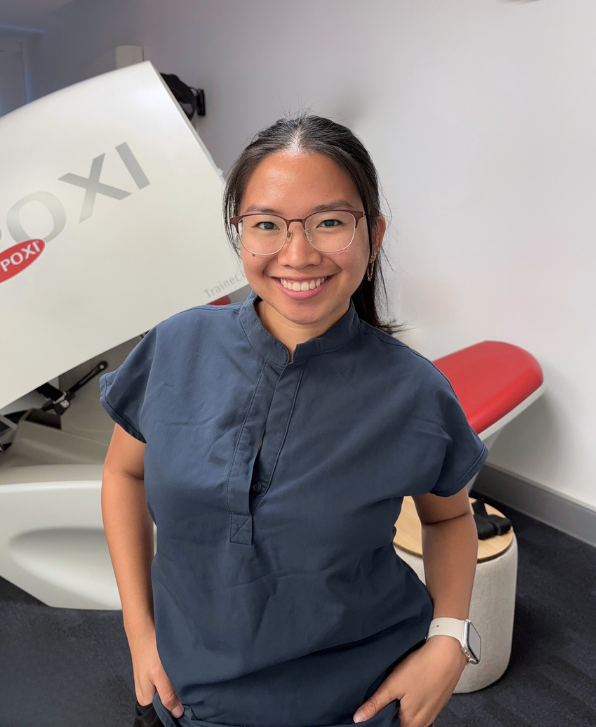 Smiling woman with glasses and a smartwatch standing in a medical office, with a large X-ray machine in the background.