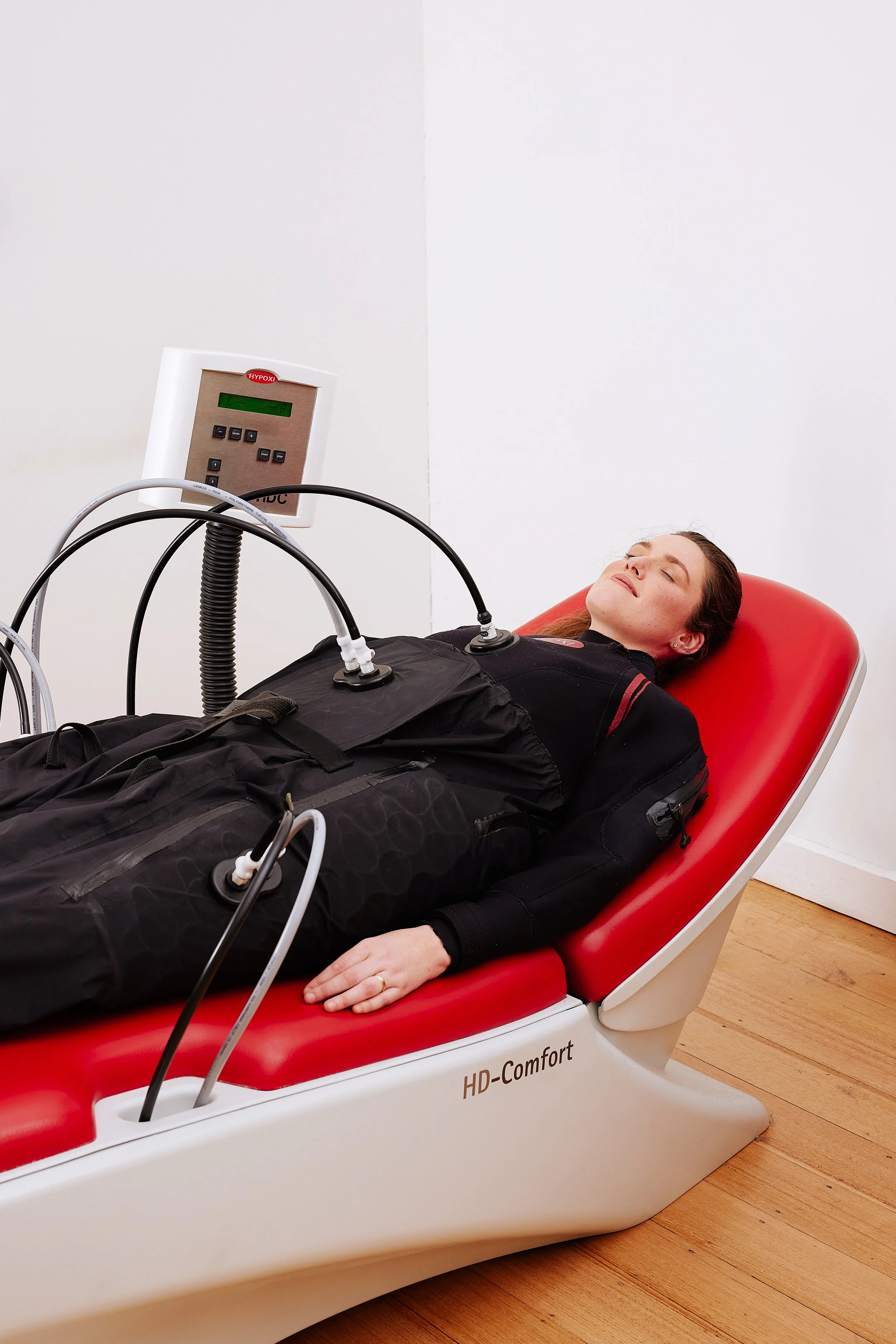 A woman lying on a red and white hyperbaric oxygen therapy chamber labeled 'HD-Comfort,' receiving oxygen treatment through multiple tubes connected to a monitor.