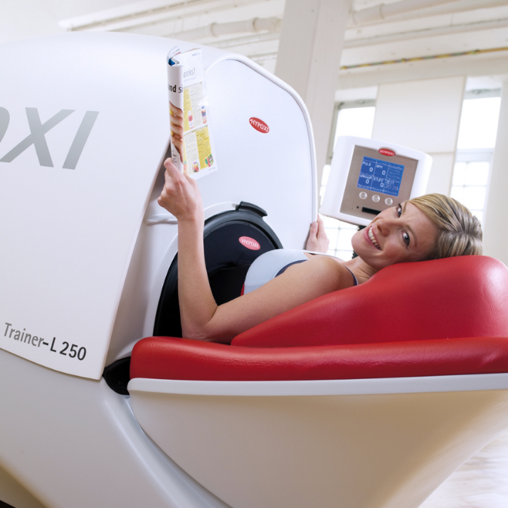 Woman lying inside a hyperbaric chamber, smiling, holding a carton, around medical equipment and monitor.