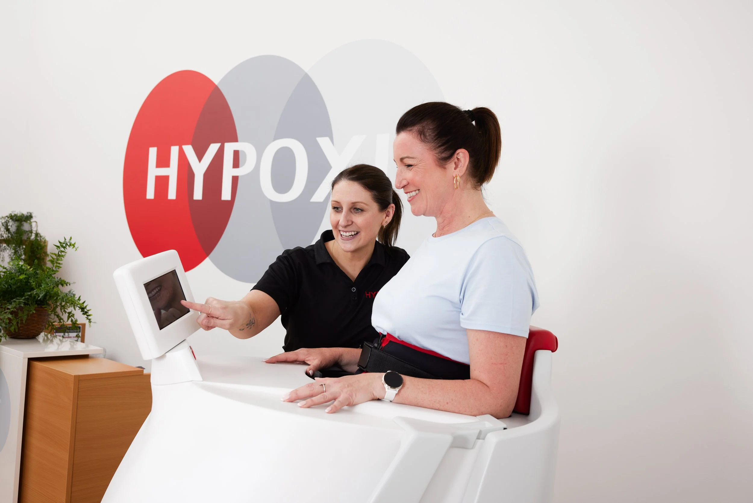 A woman sits at a medical device with a woman medical professional pointing at a screen, in front of a wall with a logo reading 'HYPOX'.