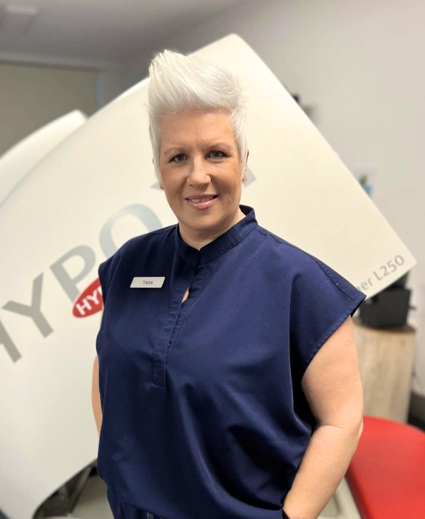 A woman with short platinum blonde hair smiling, wearing a navy blue uniform, standing in front of a large medical imaging machine.