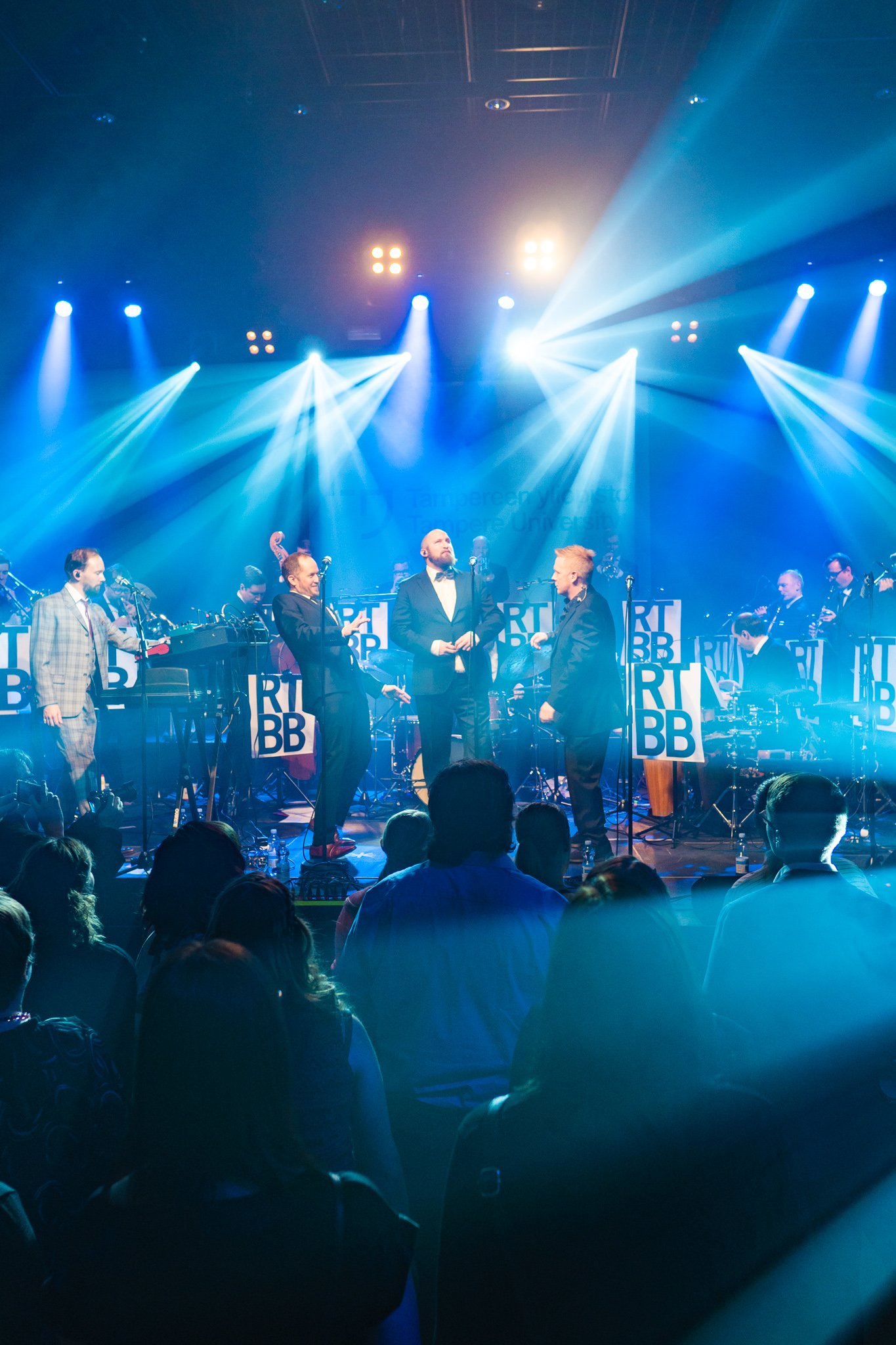 A live concert performance with musicians and singers on stage under bright blue and white lights, audience watching the show.