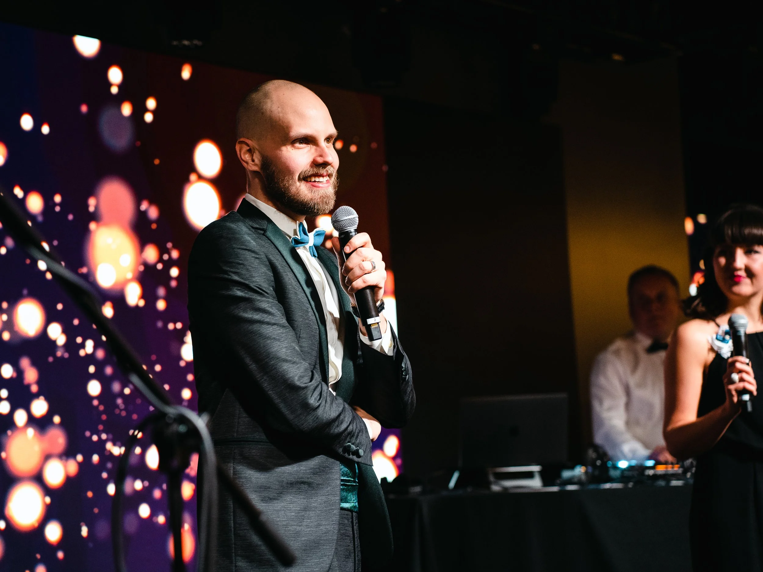 Man in tuxedo holding microphone on stage with woman and DJ in background at event.