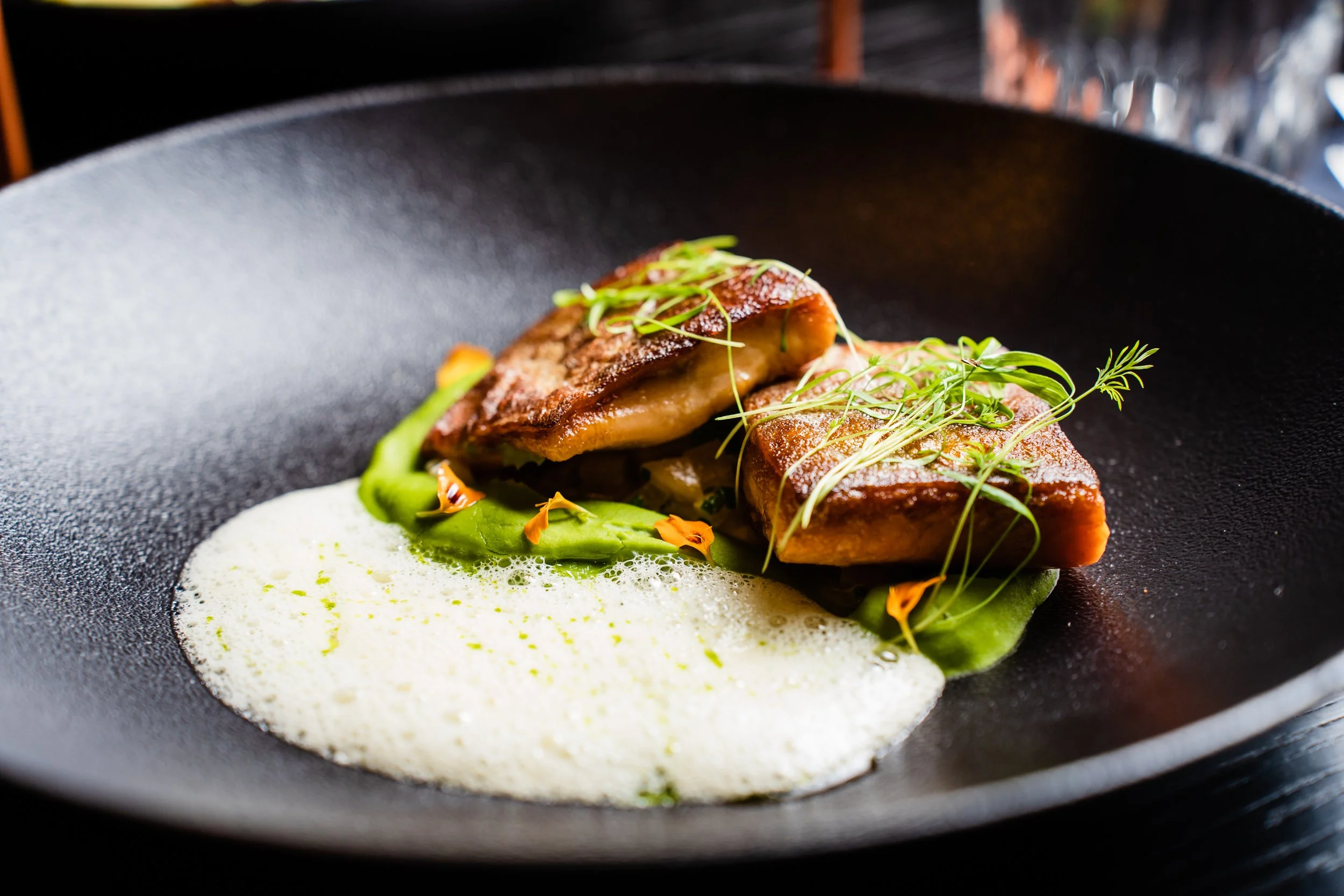 Seared pieces of fish garnished with microgreens, served on a bed of green puree and creamy foam on a black plate.